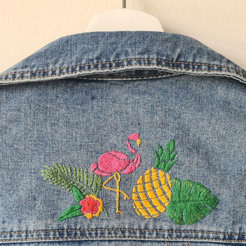 A denim jacket is on a hanger against a white wall. The back of the jacket shows a hand embroidered design of a flamingo, pineapple, flower, and monstera leaf