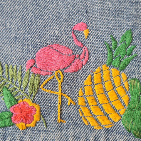 A close up of the back of a blue denim jacket. In the centre, several bright hand embroidery designs are stitched on in a collage: a flamingo, a pineapple, a monstera leaf, and a pineapple.