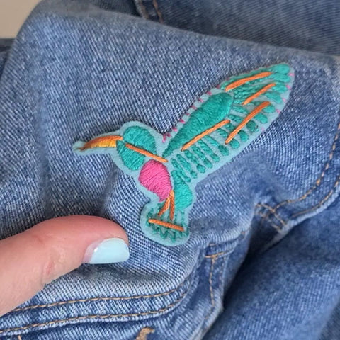 a close up of a piece of hand embroidered patch showing a hummingbird design. The patch is being attached to the front of a denim jacket.