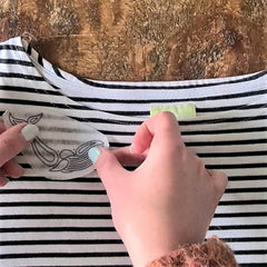 A striped t-shirt is lying on a wooden table. A pair of hands are holding a small piece of paper showing a whale design, sticking it to the shirt