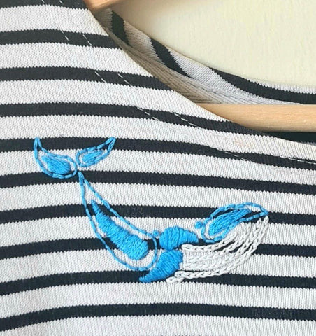 A close up of a stripy shirt, on a hanger against a white wall. Near the shirt neckline is a hand embroidered design of a whale in white and blue thread.