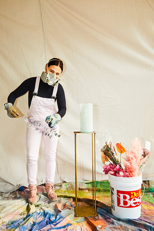 woman in pink overalls and a gas mask spray painting flowers in a white art studio