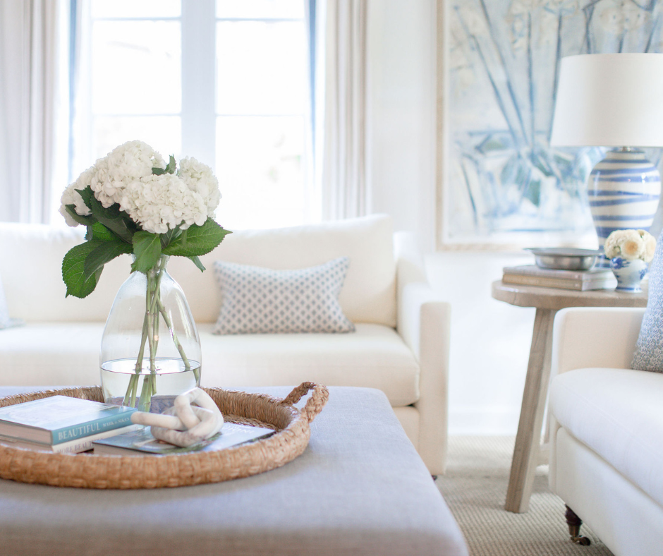 Coffee table close-up with white flowers in clear vase no top over white couch next to an illuminated window