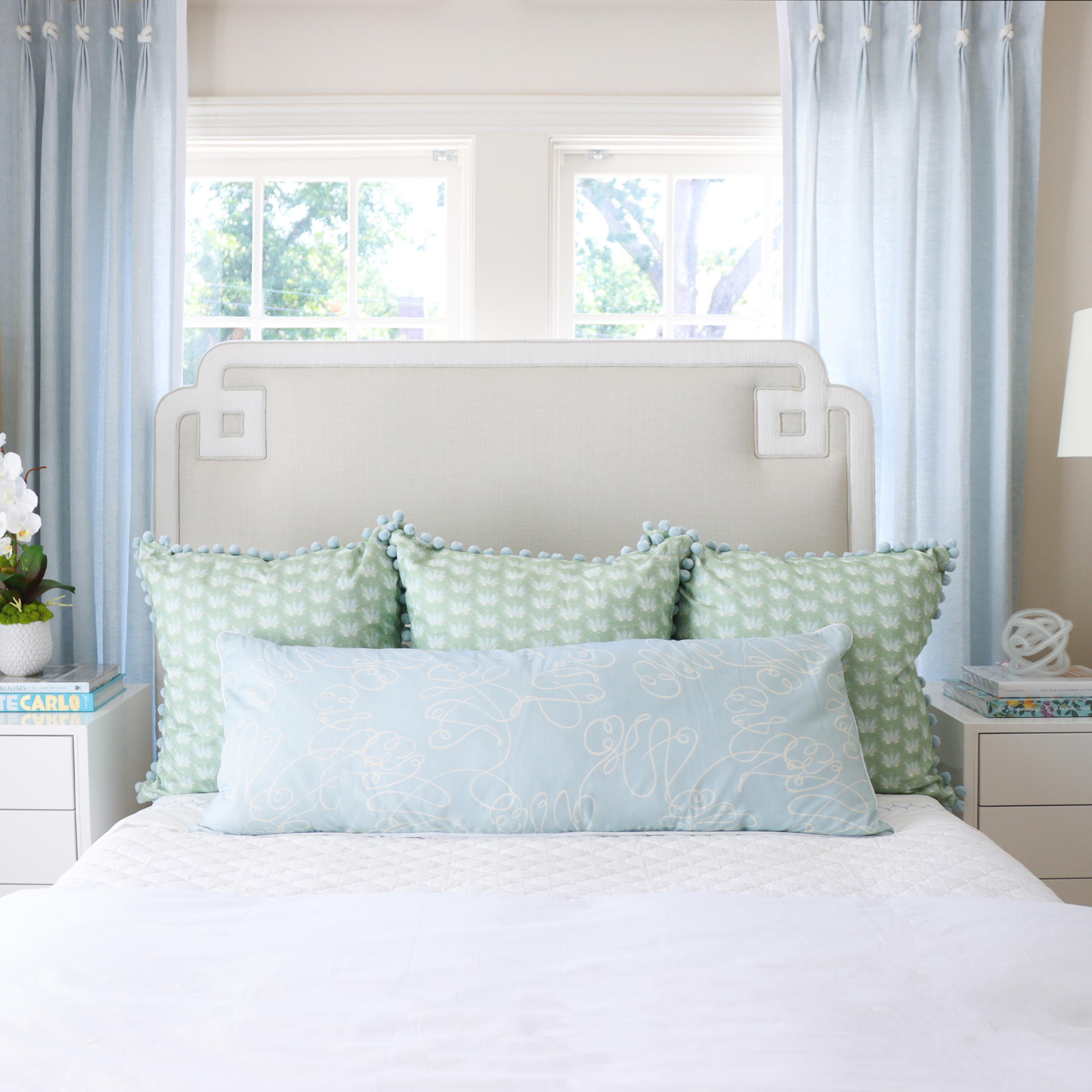 White bed close-up styled with three Blue & Green Floral Drop Repeat Printed Pillows and one Powder Blue Abstract Printed Lumbar pillow in front of an illuminated window with one white night stand on each side