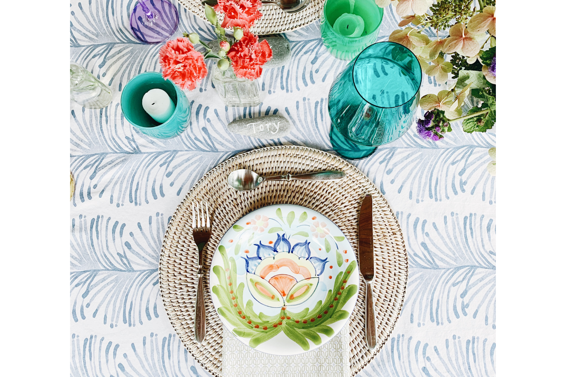 overhead shot of place setting with sky blue botanical tablecloth and moss green geometric napkins paired with rattan placemat, green wine glass and floral dinner plate