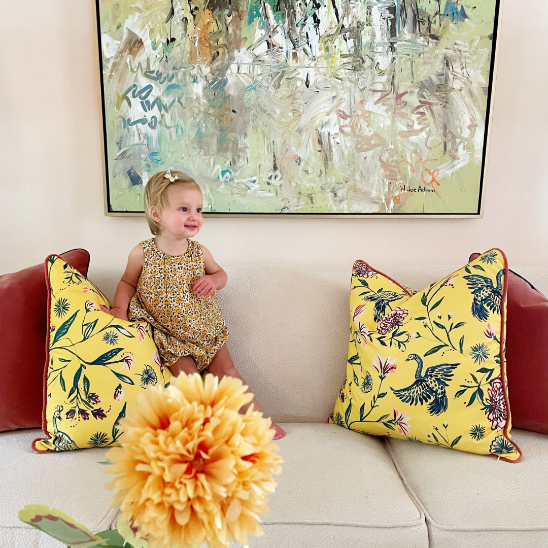 Blonde baby standing on couch in between two coral velvet custom pillows and two Yellow Chinoiserie custom pillows with a painting hung on wall
