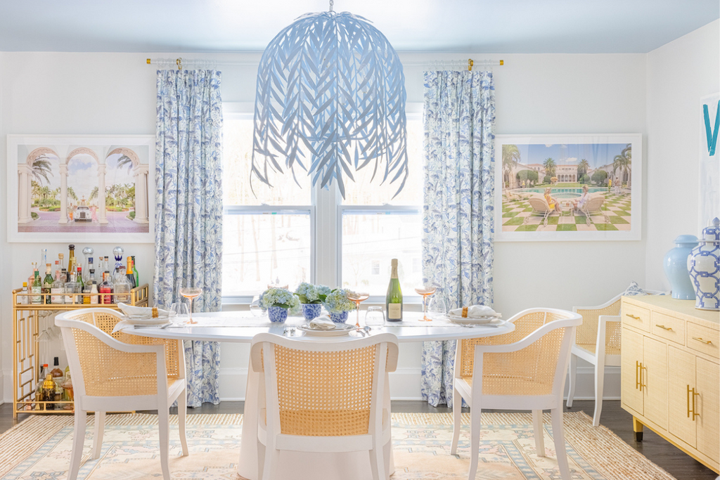 Bright dining room with blue tiger chinoiserie curtains hanging on a rod over a double window styled with a white oval table and white cane chairs and blue whimsical chandelier