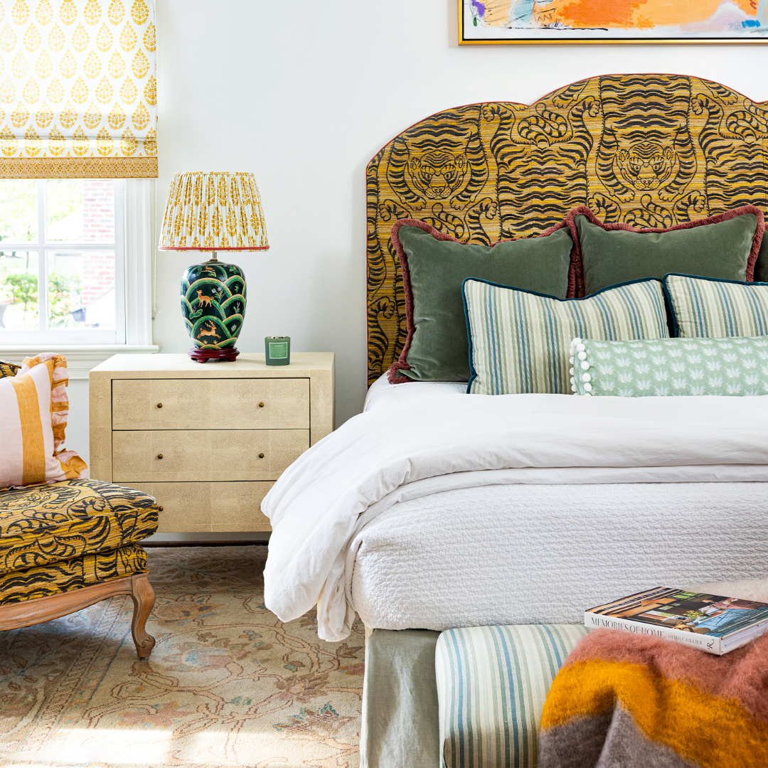 Bedroom with a wooden nightstand with a lamp and candle on top next to a white bed styled with a tiger printed headboard and two fern green velvet custom pillows with two striped lumbar pillows in front and a Blue & Green Floral Drop Repeat custom bolster with white pom poms
