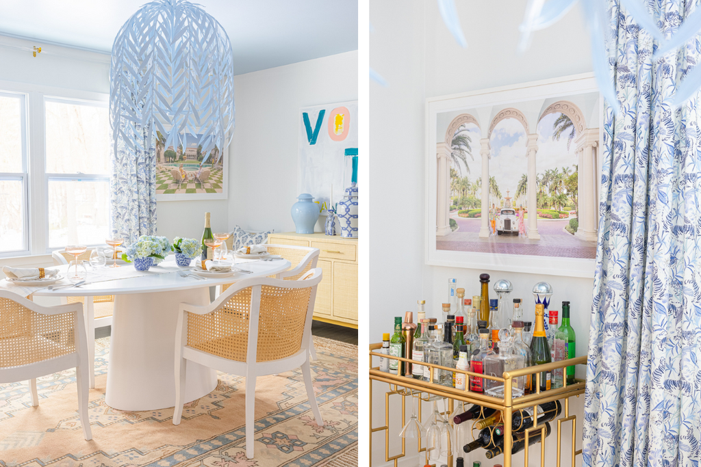 Bright dining room with blue tiger chinoiserie curtains hanging on a rod over a double window styled with a white oval table and white cane chairs and blue whimsical chandelier next to a bart cart with palm beach colorful artwork