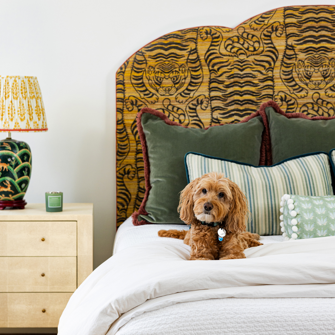 Brown dog on top of white bed styled with two fern green velvet custom pillows with two striped lumbar pillows in front and a Blue & Green Floral Drop Repeat custom bolster with white pom poms next to wooden nightstand