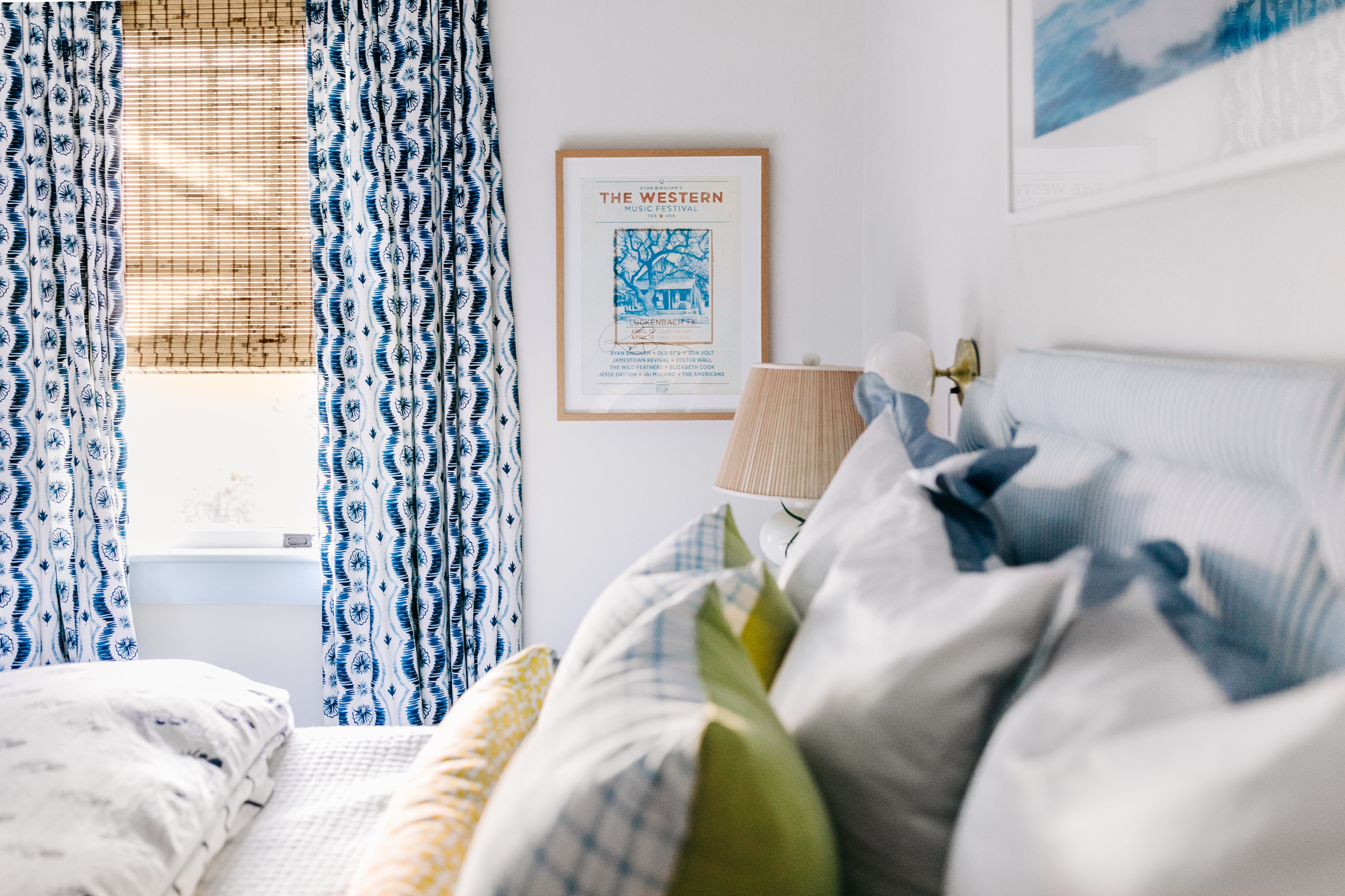 Blue ikat custom curtains hanging on a window with woven roman shades in a white bedroom with a blue striped headboard styled with colorful pillows