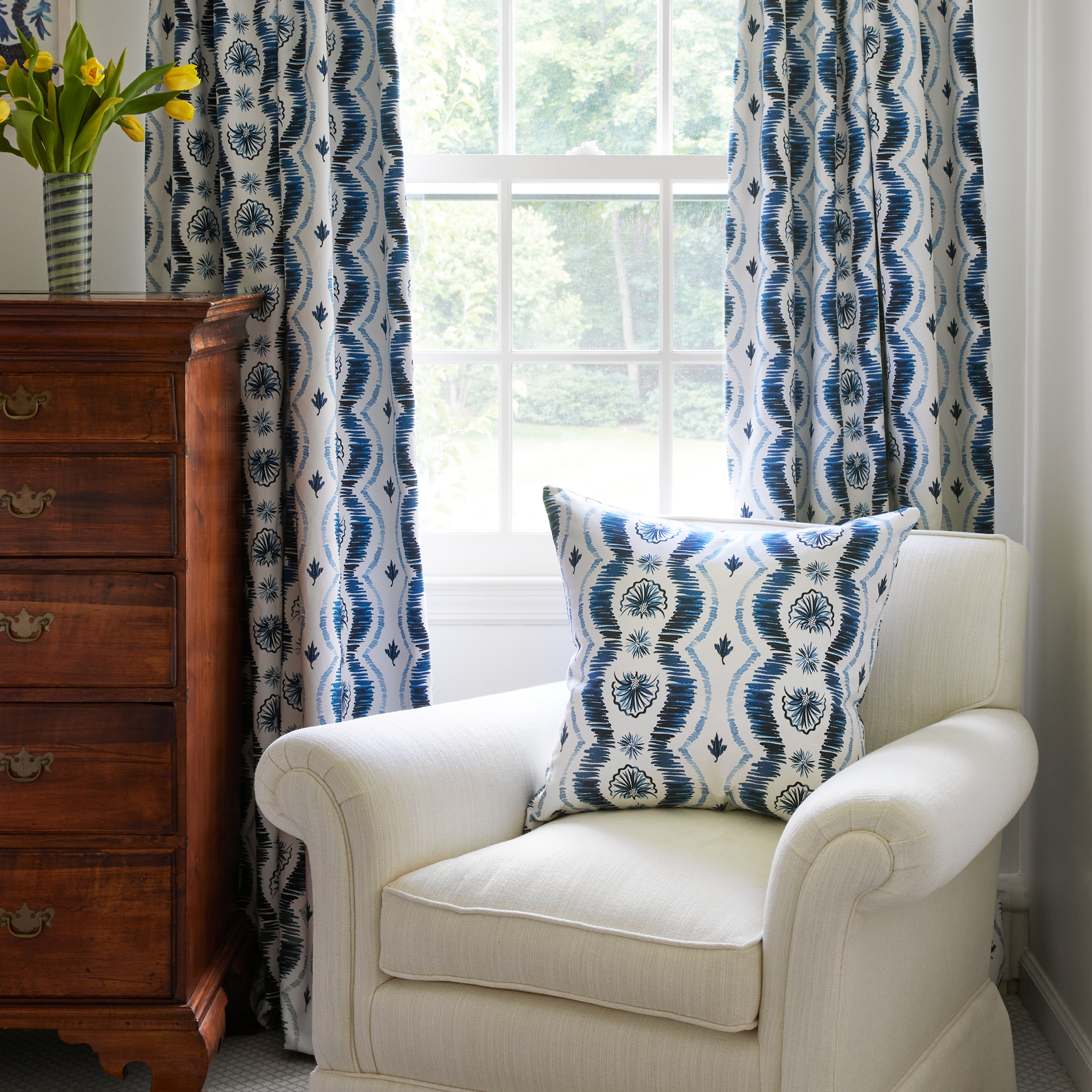 Blue ikat custom curtain hanging on a rod in front of a window next to a tall wooden chest and a white upholstered chair styled with a blue ikat custom pillow