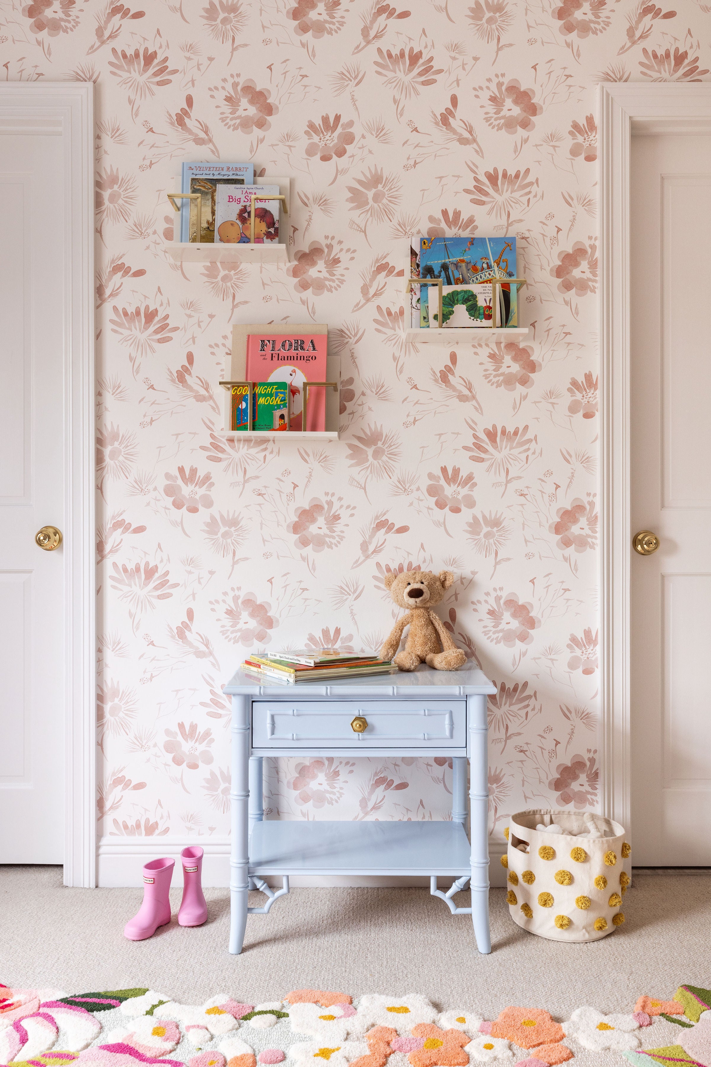 Purple nightstand between two doors in a nursery with pink floral wallpaper and acrylic floating shelves styled with kids books