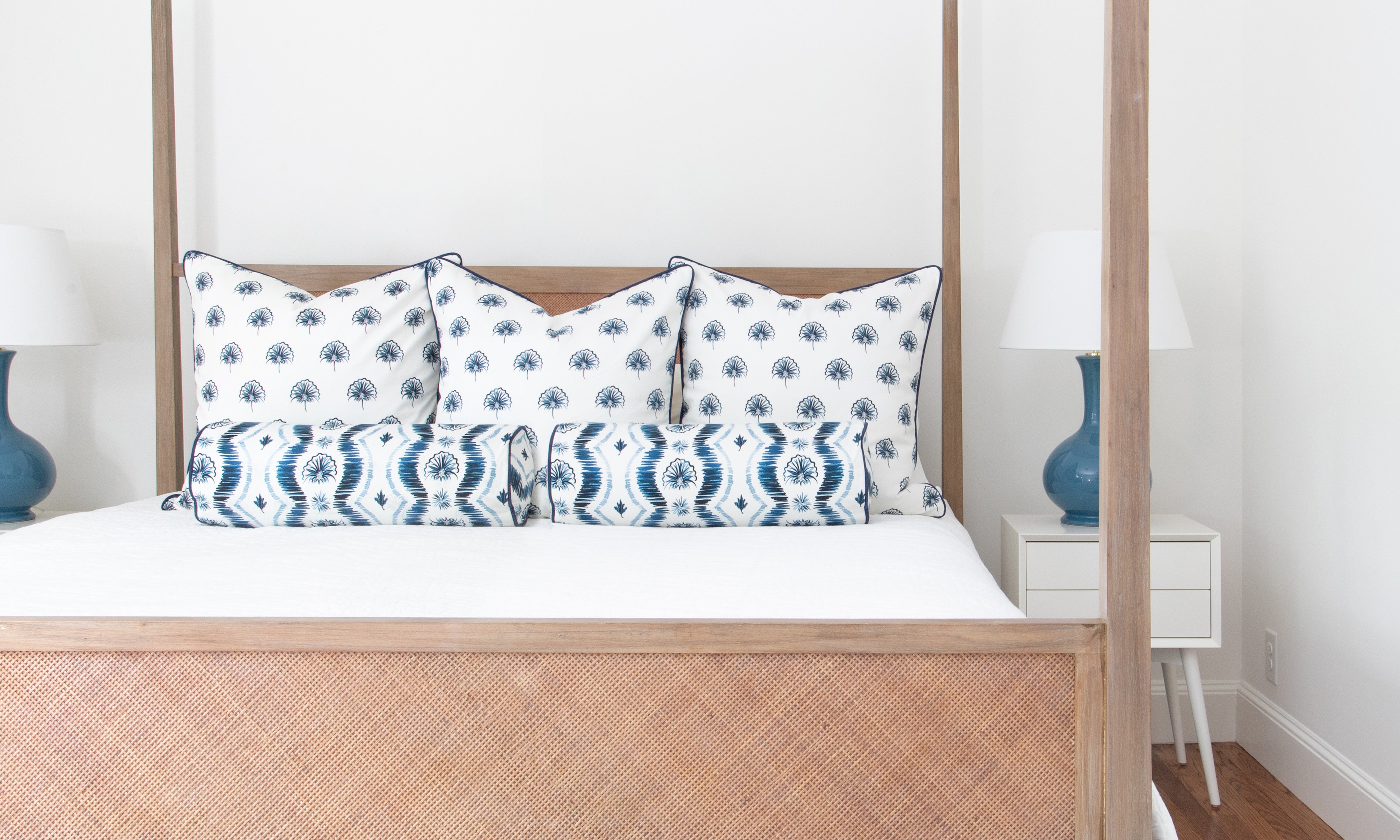 Blue ikat striped custom pillows and floral navy custom pillows styled on a light wood bed with white sheets in a room with white walls.