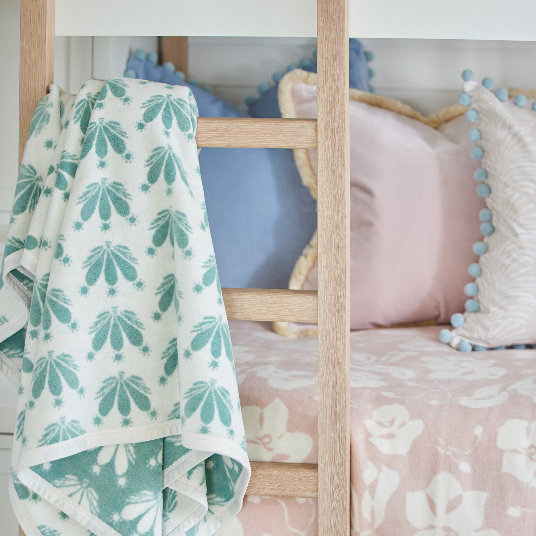 Blue and green floral blanket hanging on a wooden latter attached to a bunk bed with a pink floral blanket covering the bottom bunk styled with blue and pink pillows