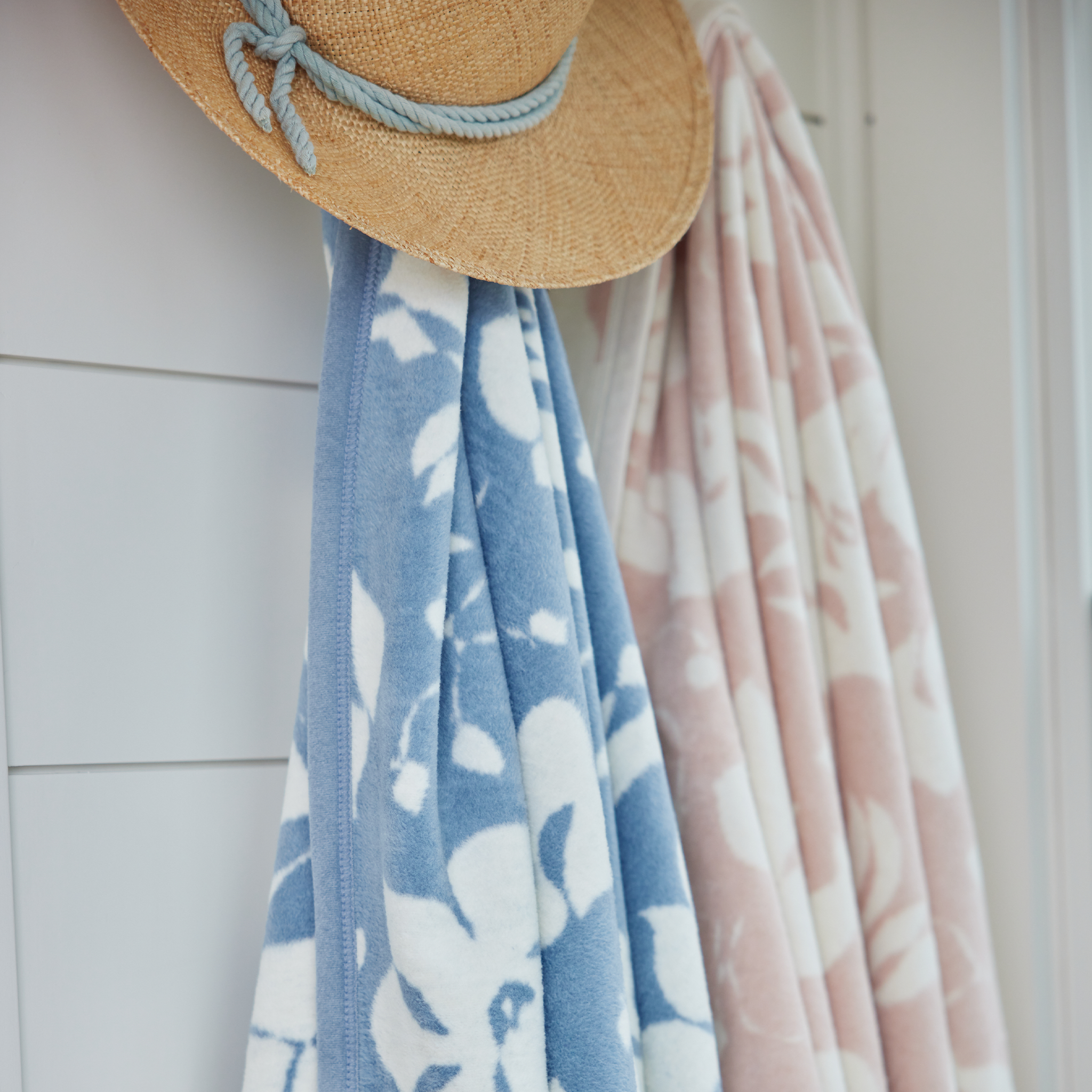 Close up of one blue and white floral blanket and one pink and white floral blanket hanging on hooks on a white wall styled with a straw sun hat