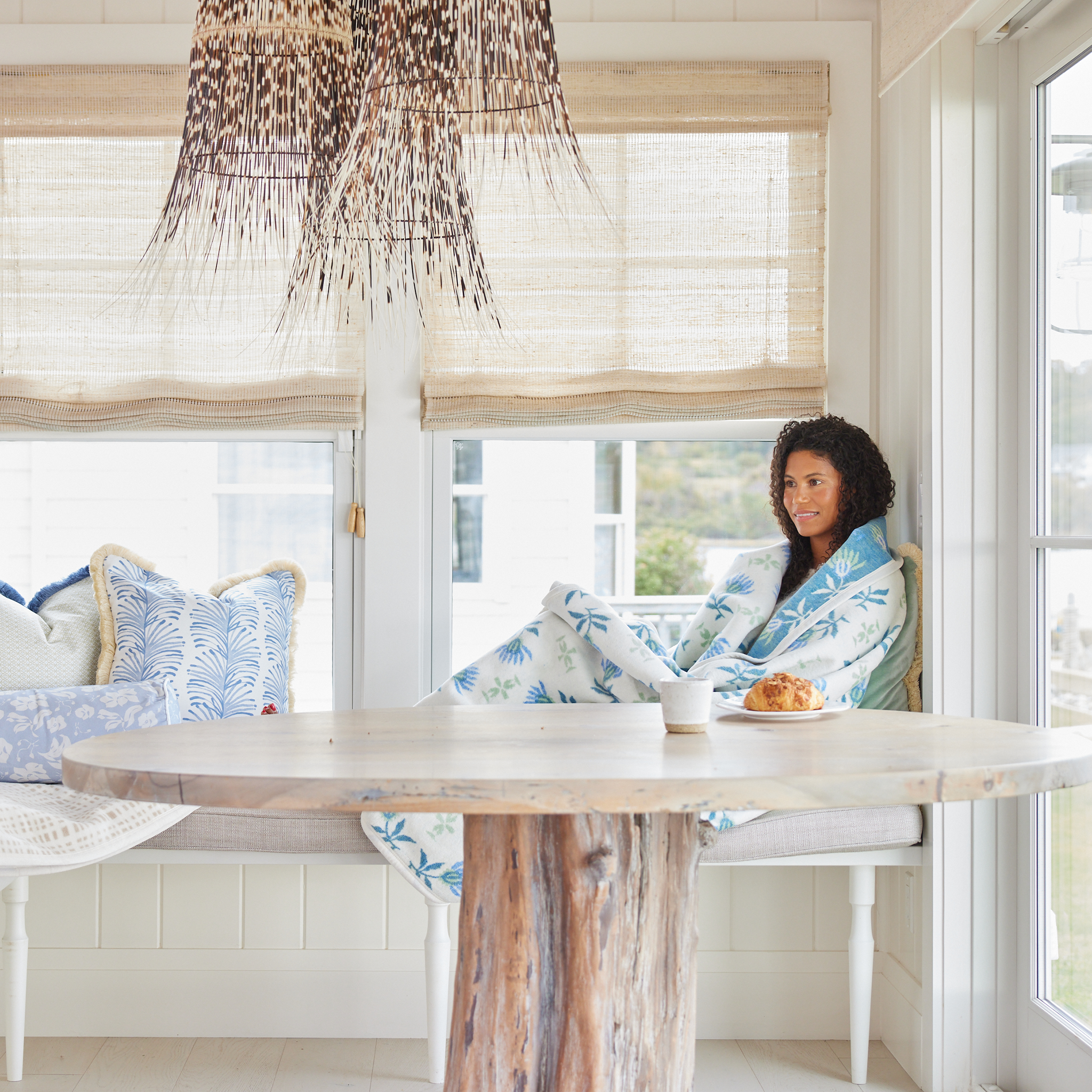 Brunette woman sitting on a kitchen bench in a coastal-inspired home wrapped up in a blue floral blanket laying on a green velvet pillow behind a wooden table