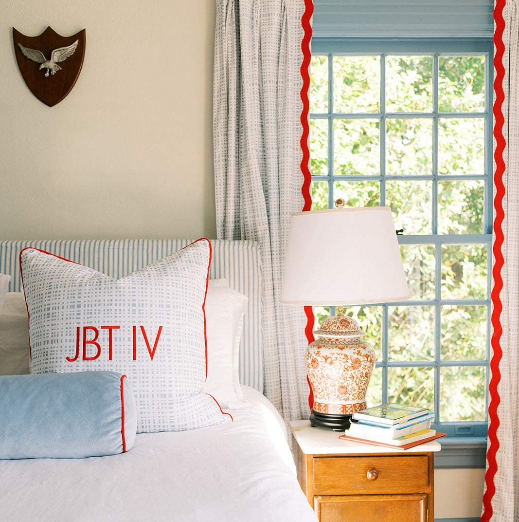 Close up of a bed with blue gingham pillow piped in red and a sky velvet bolster piped in red next to a window with blue gingham curtains and red rick rack trim