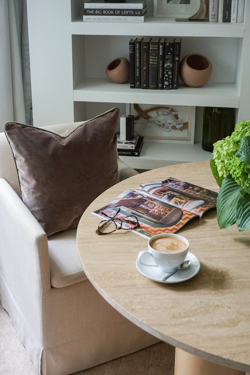 White sofa chair styled with a Brown Velvet Custom Pillow next to a circular table with a cup of coffee, glasses, magazine, and flowers in clear vase on top