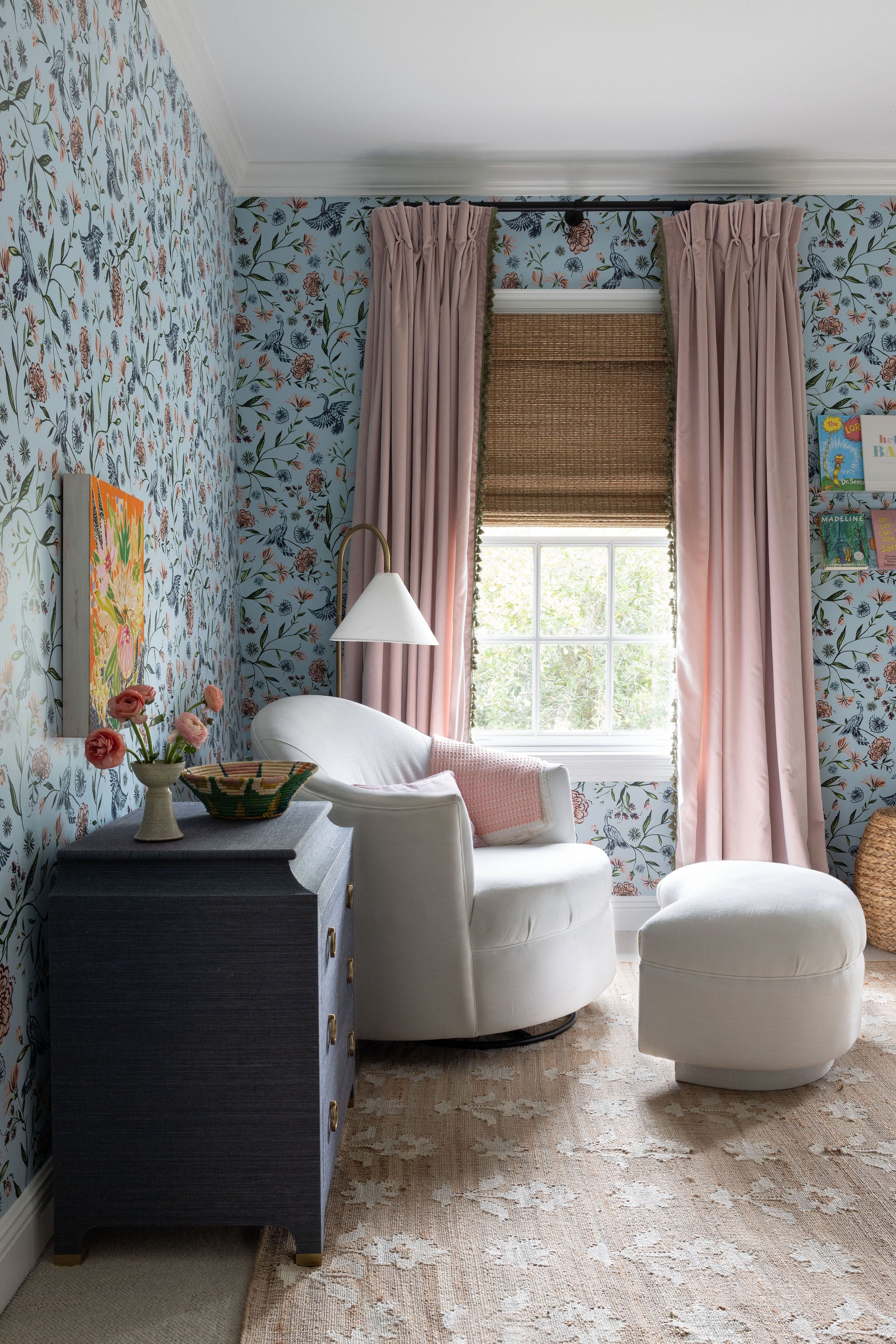 Corner of a nursery with blue chinoiserie wallpaper paired with pink velvet custom curtains with green tassel trim hanging on a rod in front of a window next to a white upholstered chair and pouf on a tan rug