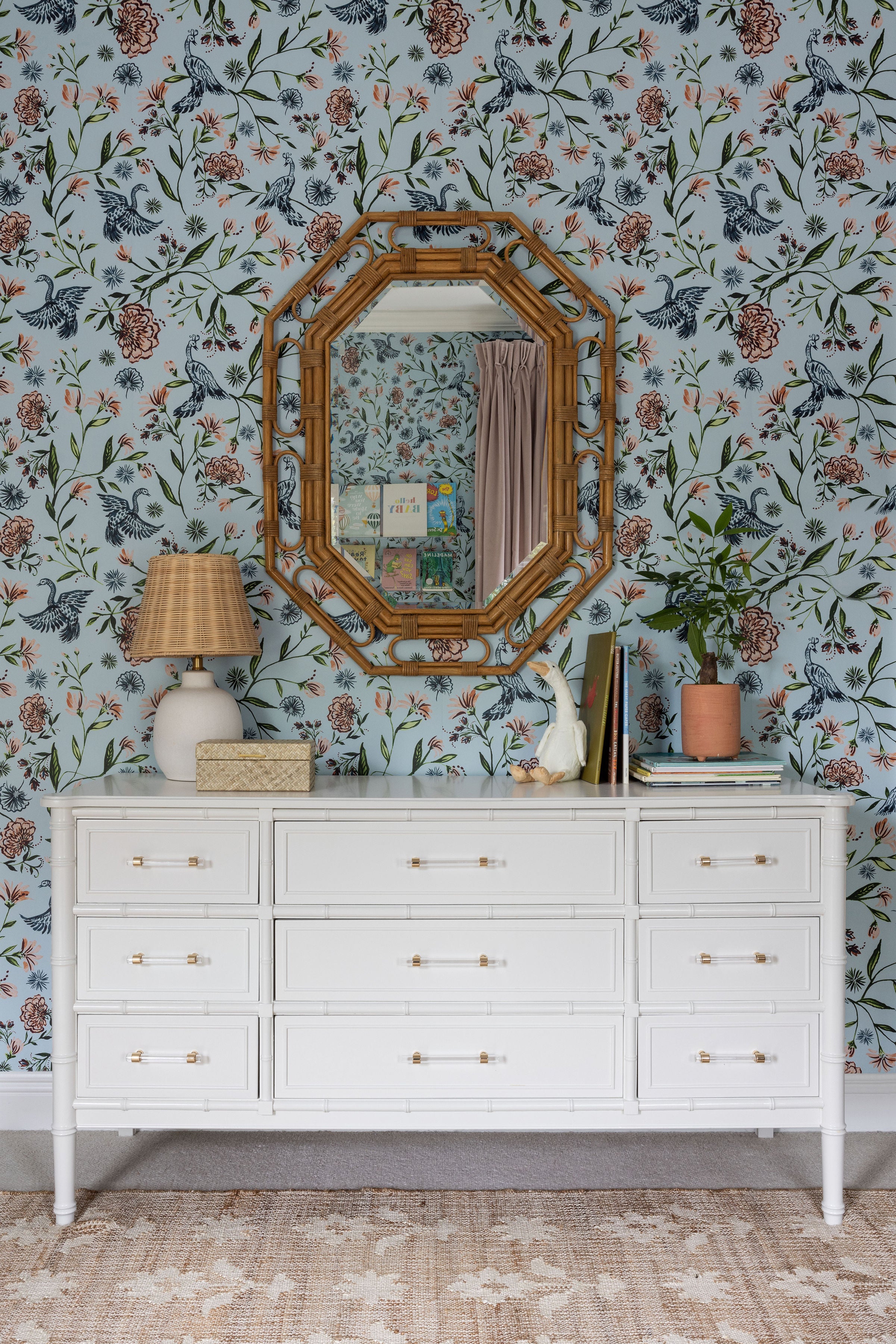 Nursery with blue chinoiserie wallpaper styled with a tan rug, white dresser and rattan mirror