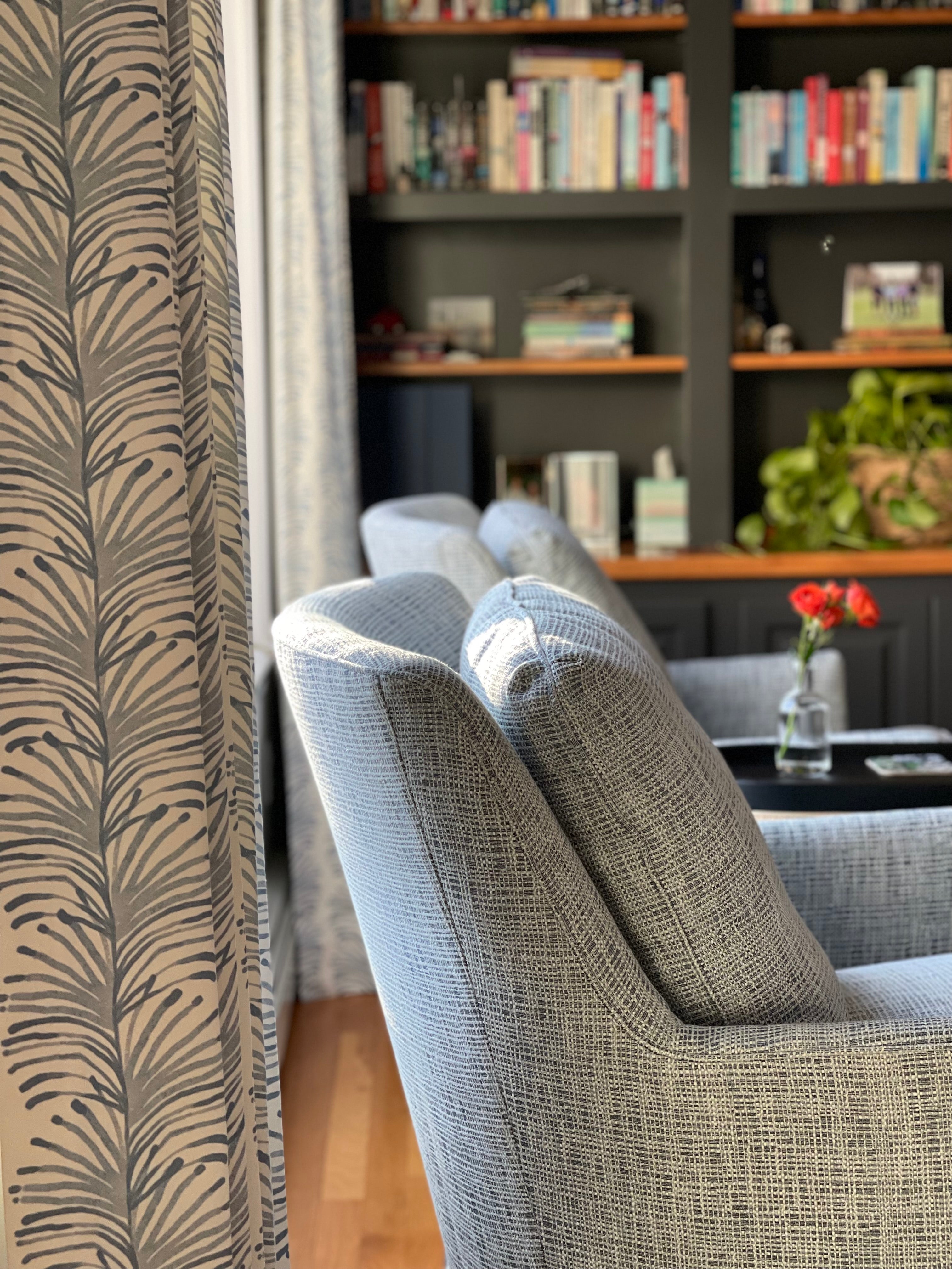 Sky blue custom curtain panel hanging on a window behind a blue upholstered chair in a living room with a navy bookcase