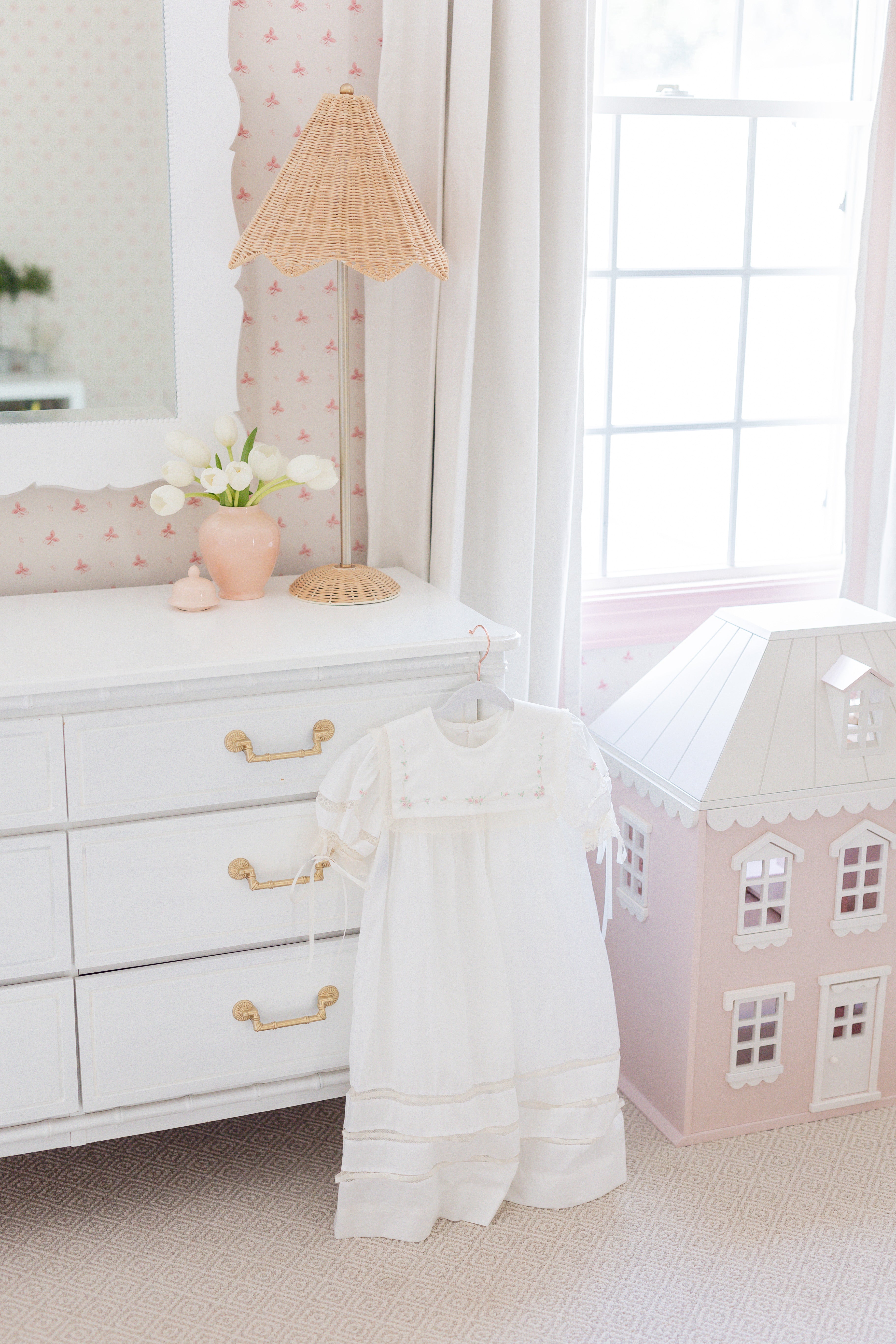 Pink and white nursery with white dresser and a white baby gown hanging on the side next to a pink dollhouse and natural white curtains