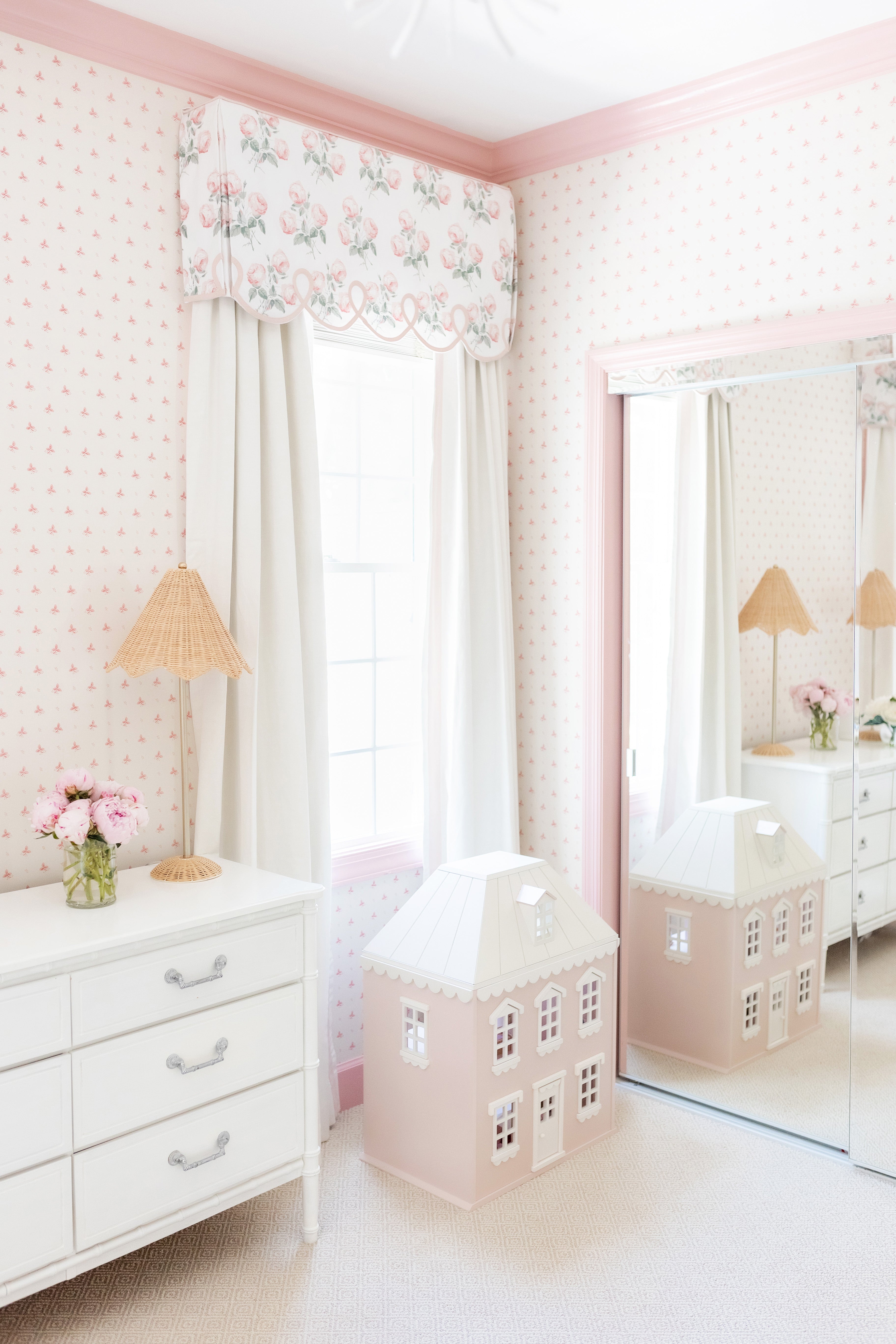 Corner of a nursery with pink and white floral wallpaper paired with natural white curtains hanging on a window with a pink and green floral valence and a white dresser and pink dollhouse