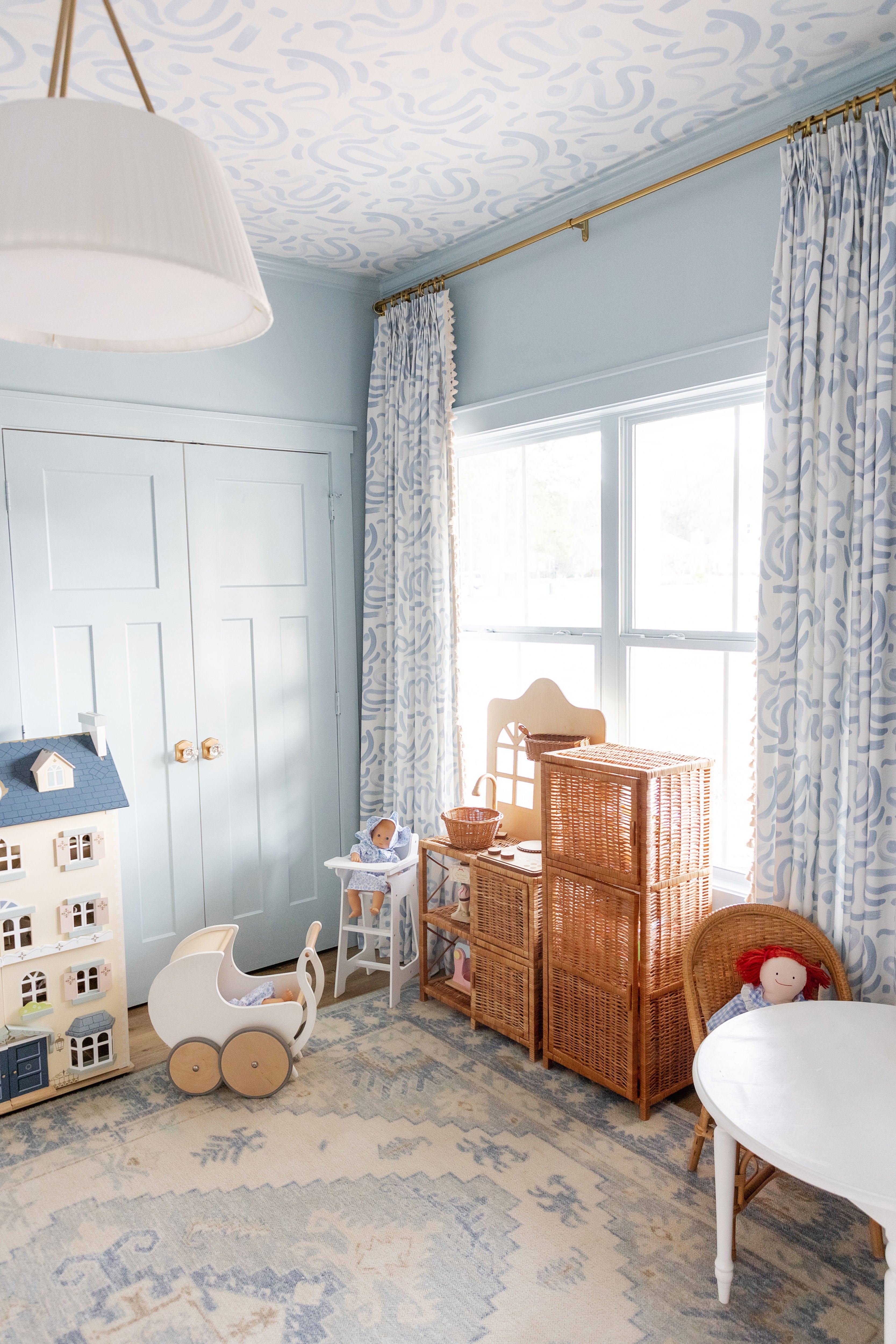 Blue painted playroom styled with rattan shelves and a blue and white rug and two sky blue whimsical curtains hanging on a rod in front of a window styled with matching sky blue whimsical wallpaper on the ceiling