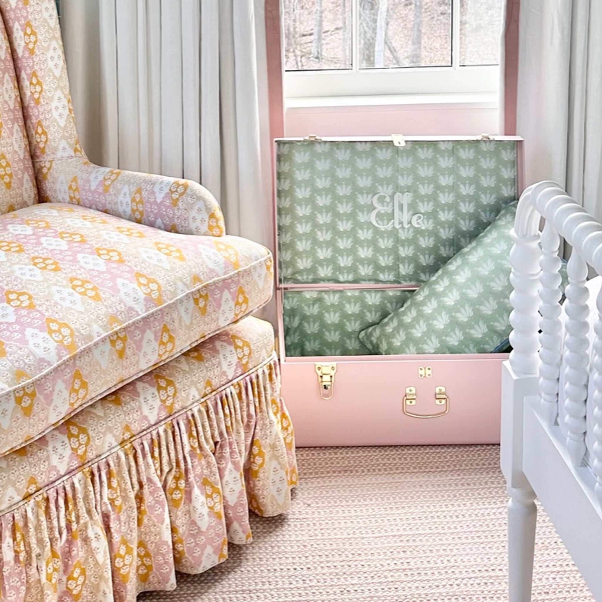 Pink keepsake trunk lined with blue and green floral fabric below a window with white curtains and next to a pink and orange upholstered chair