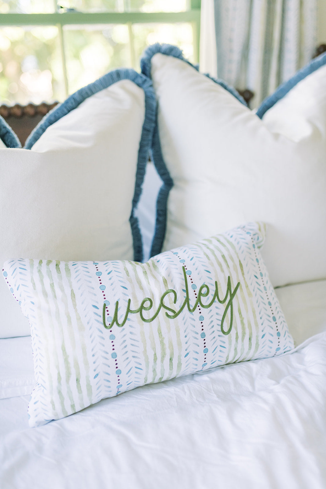 Blue and green striped embroidered pillow in front of two white euro shams with blue fringe on a bed with white sheets