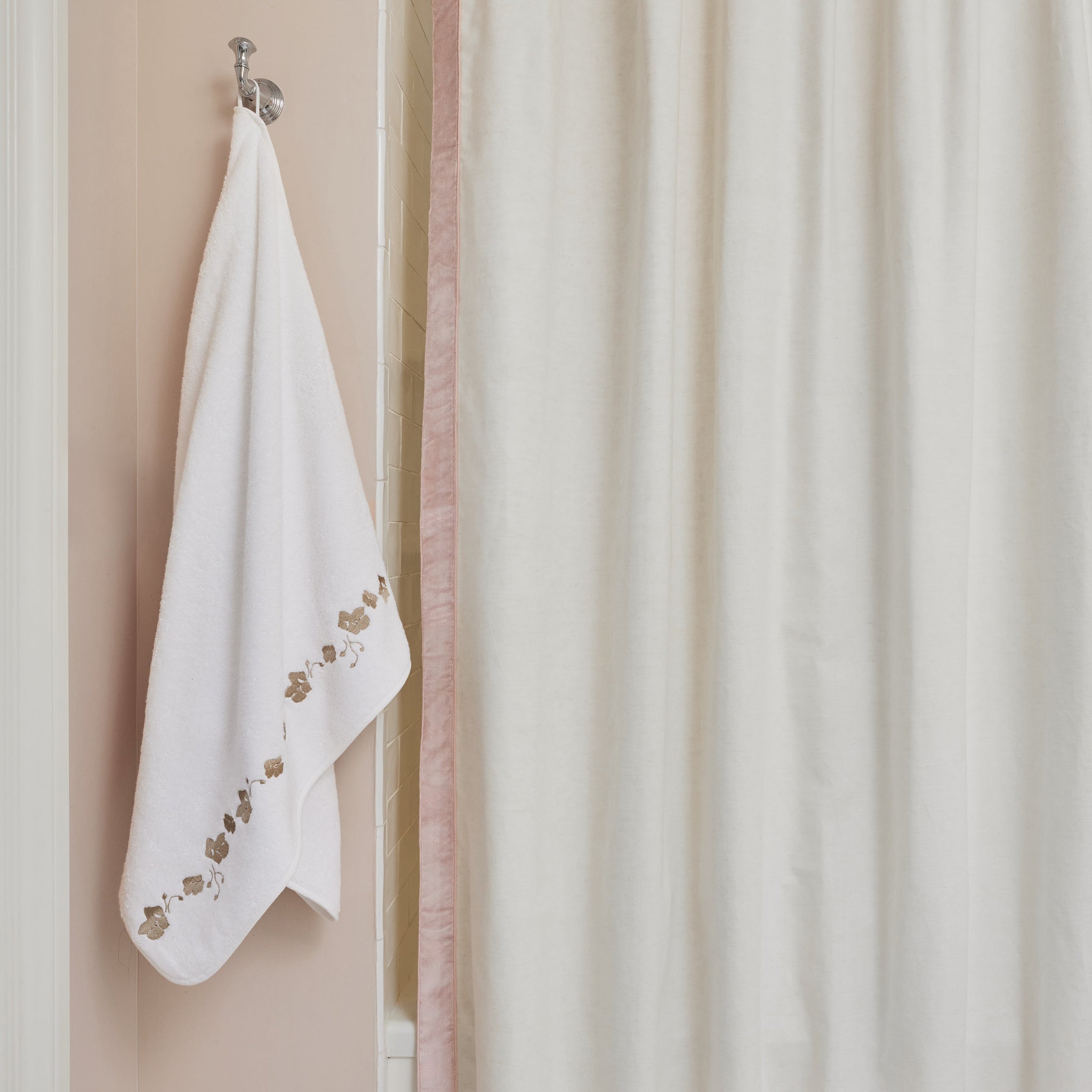 White towel with sand floral embroidery hanging on a hook next to a white shower curtain hanging on a tub in a bathroom with light pink walls