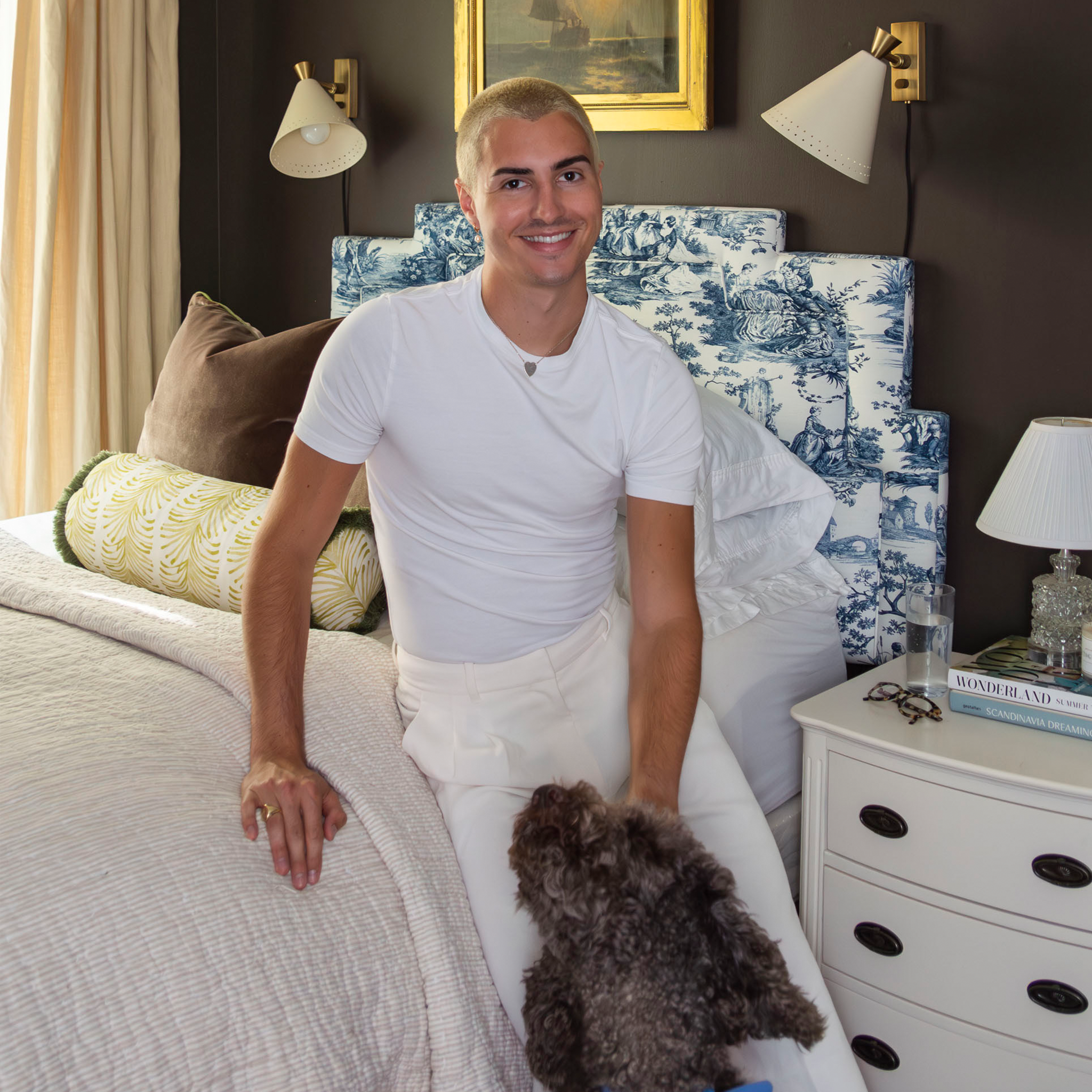 Blonde man wearing all white sitting on the corner of a white bed with one hand on a brown dog next to a dresser