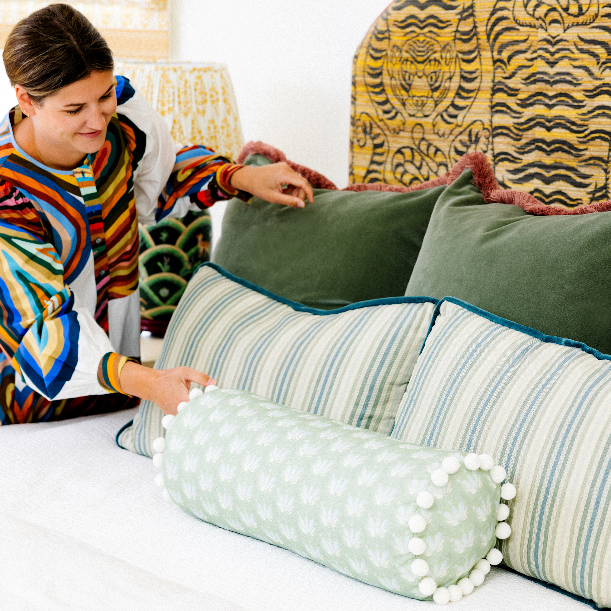 Brunette woman holding one fern green velvet custom pillow next to one other with two striped lumbar pillows in front and a Blue & Green Floral Drop Repeat custom bolster with white pom poms over a tiger printed headboard