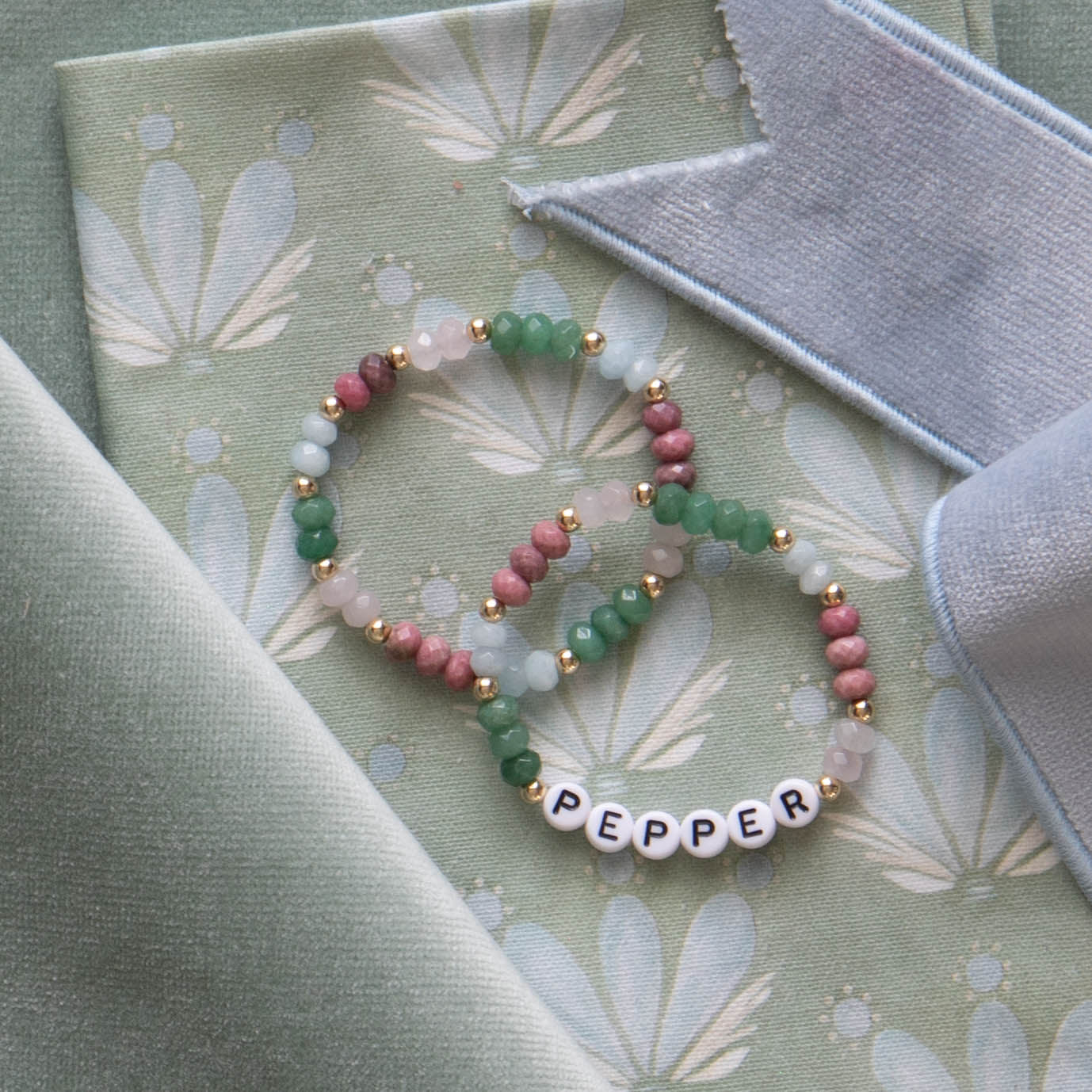 Green white and pink beaded bracelets on top of Blue & Green Floral Drop Repeat cotton fabric and green velvet fabric and sky blue velvet band