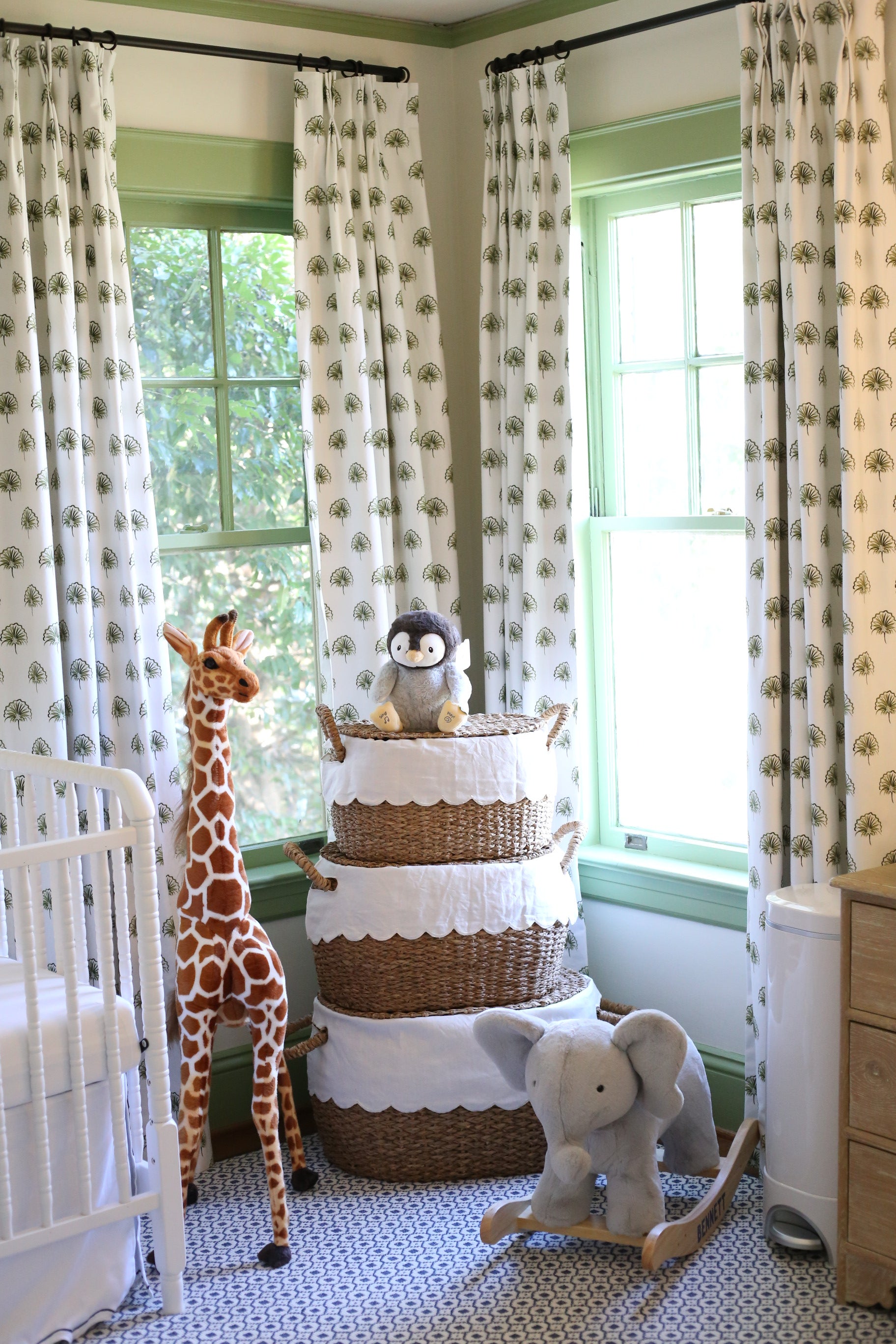 Corner of a green nursery with green floral curtains hanging on 2 windows behind stacked rattan baskets and a stuffed animal giraffe