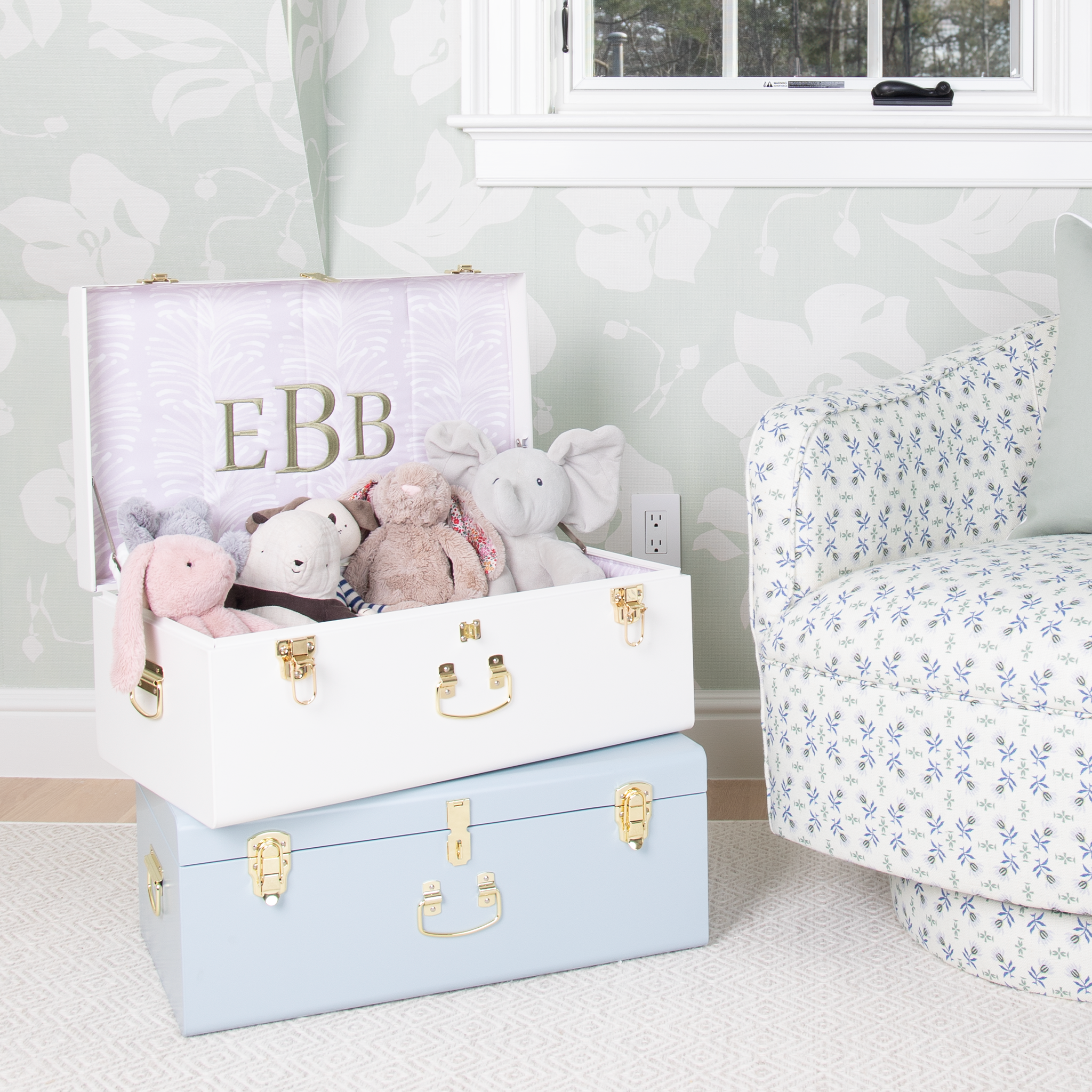 Two white and blue keepsake trunks stacked on top of each other in a room with mint floral wallapper and and a blue and green floral patterned swivel chair