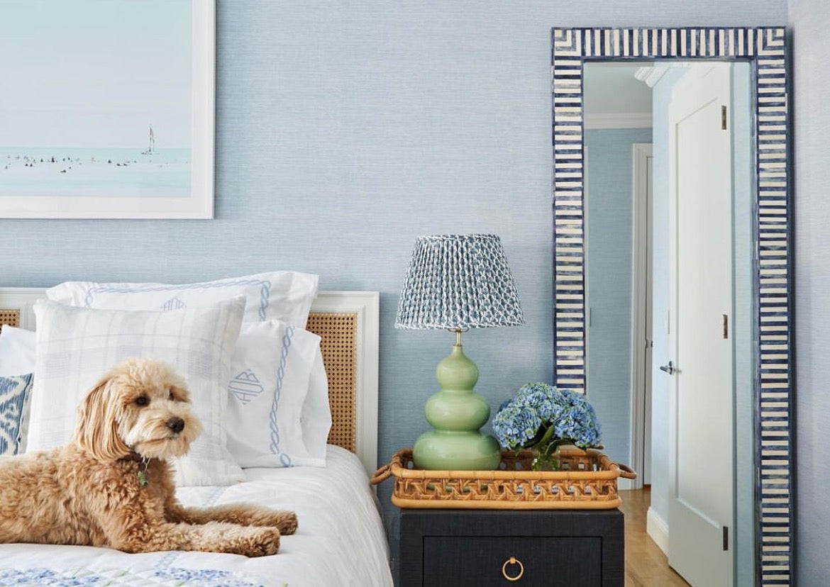 Dog laying on white bed next to wooden nightstand with green lamp and flowers on top