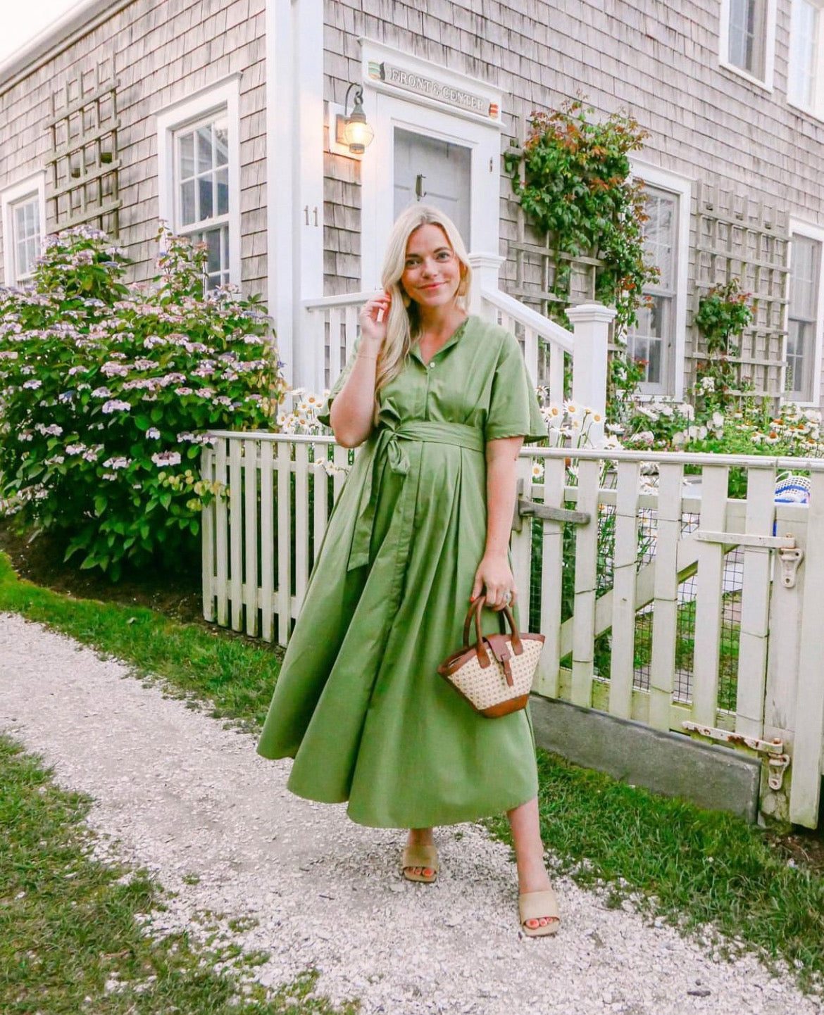 Blonde woman wearing green long dress standing in front of a white fenced house