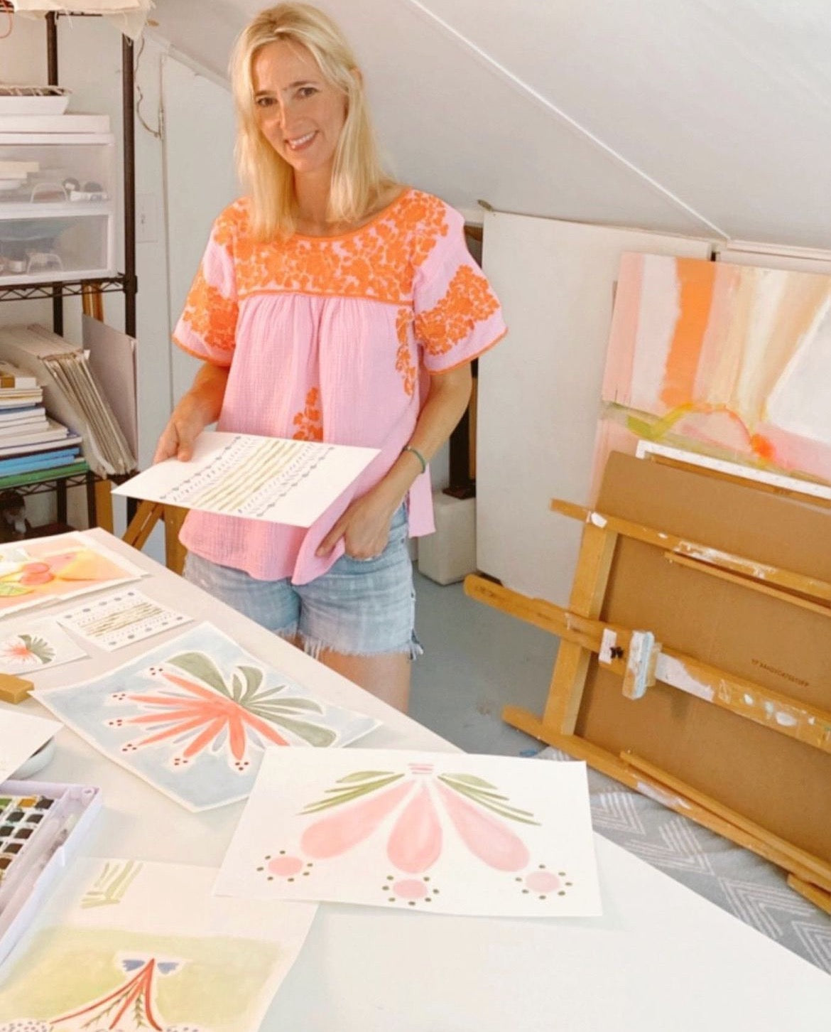 Blonde woman in pink and orange top and jean shorts standing in her art studio looking at floral watercolor artwork