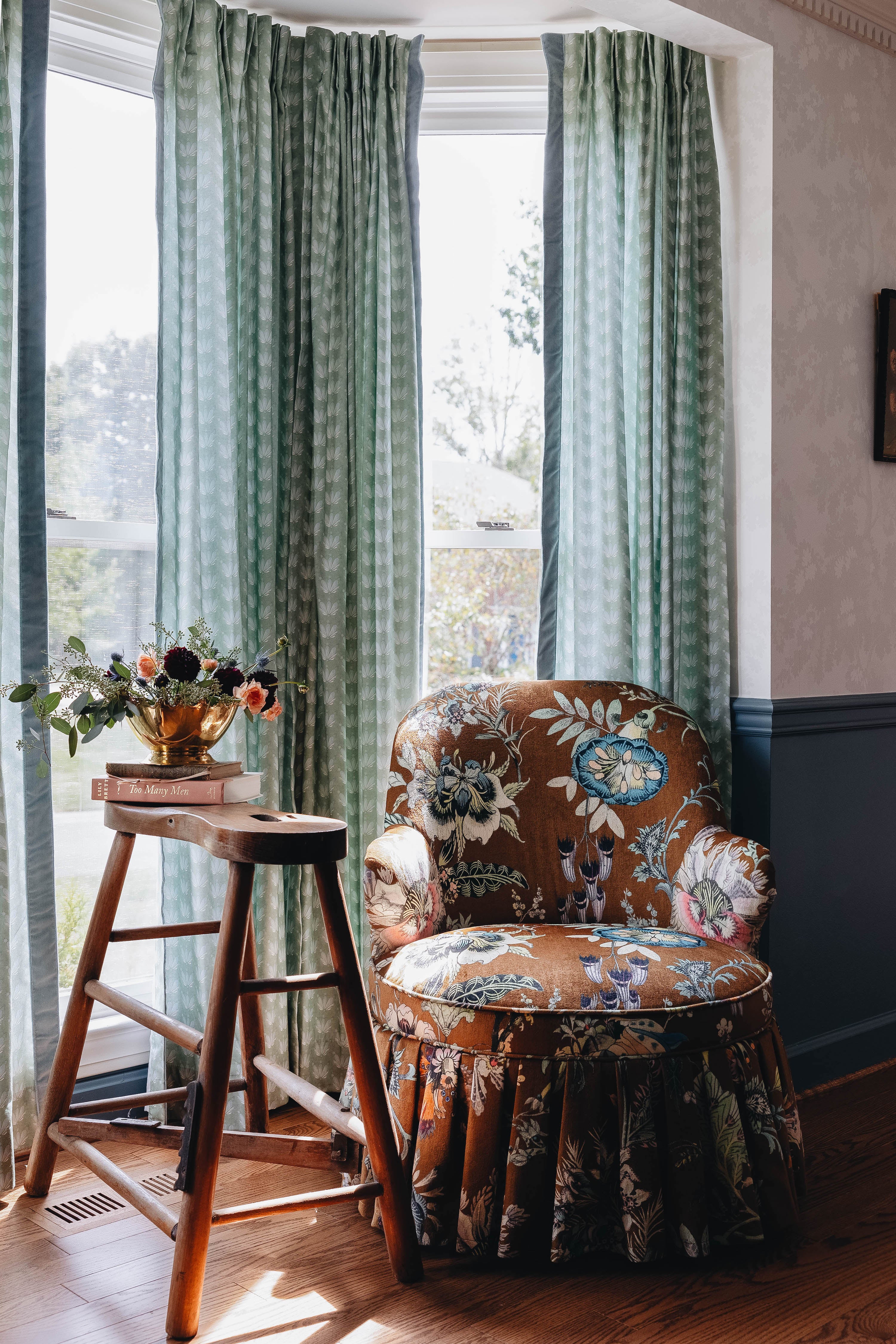 Brown and green patterned chair in front of coastal inspired green and blue curtain panels styled with a wooden stool and burgundy and pink flowers