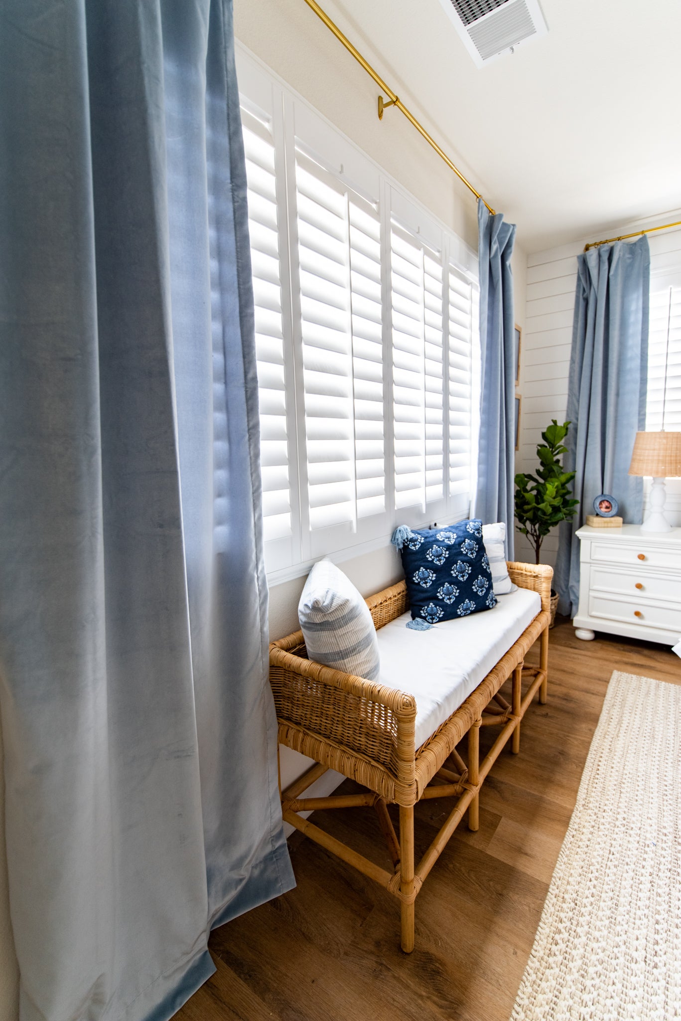 Sky velvet custom curtains hanging on a gold rod over a large window with white shutters and styled with a rattan bench below