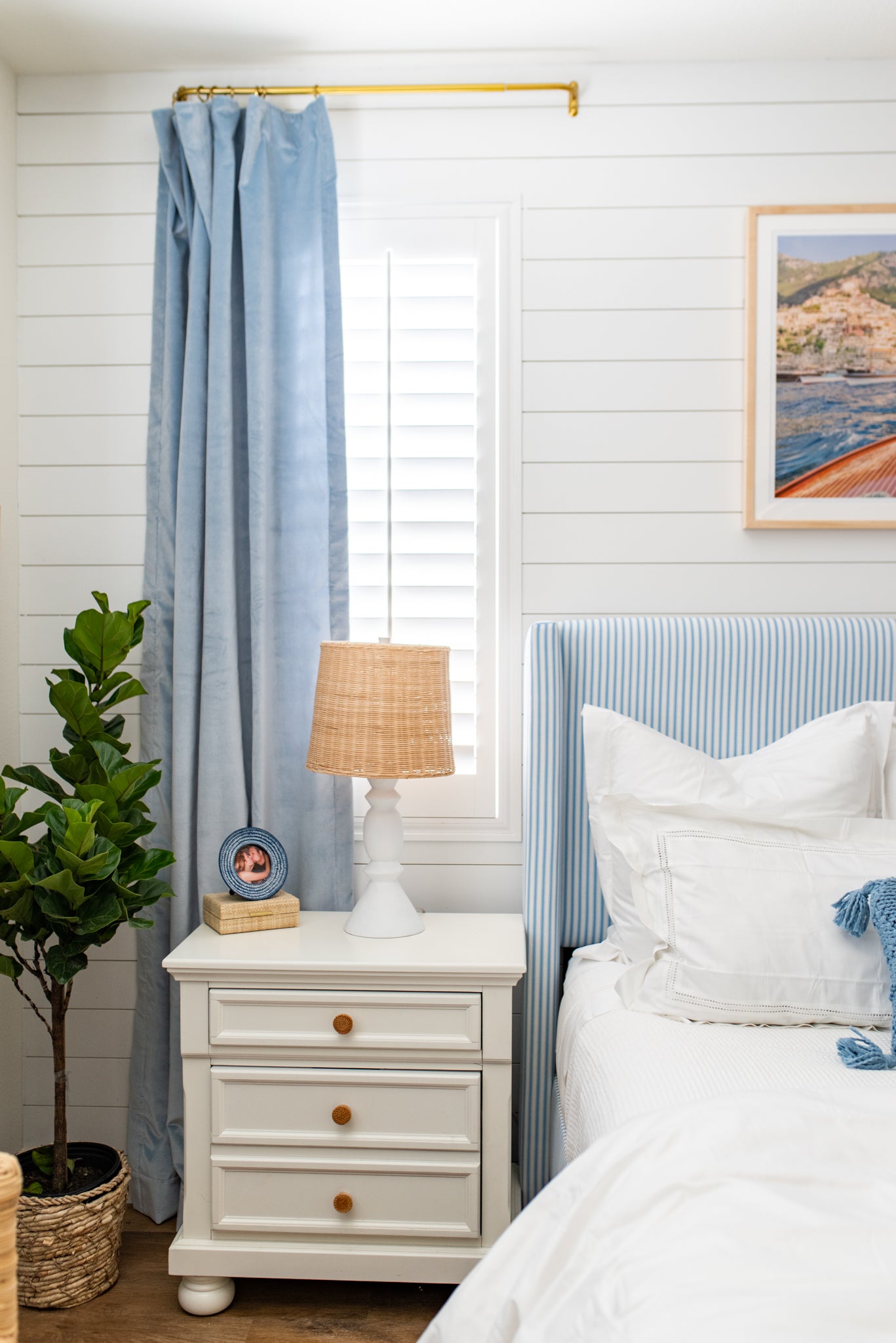 Close up of a coastal inspired bedroom with sky blue velvet curtains hanging on a rod in front of a window styled next to a blue and white striped headboard with white sheets and a white dresser