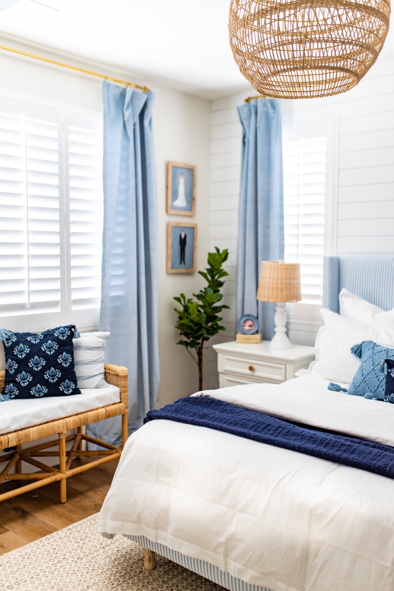 Corner of a coastal inspired blue and white bedroom with sky blue velvet curtains hanging on two windows styled next to a blue and white striped headboard with white sheets and a white nightstand and rattan bench