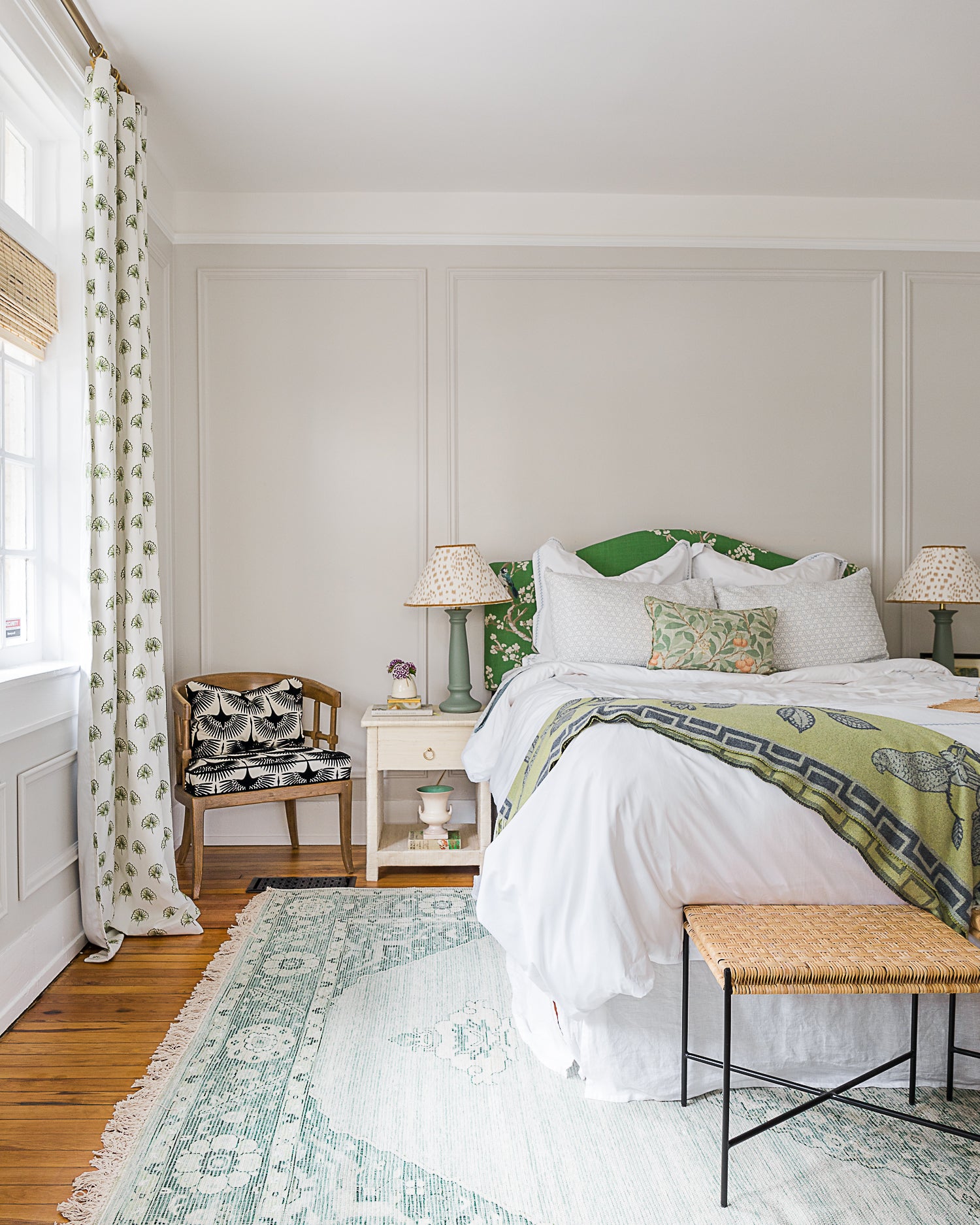 Bedroom with a white bed over rug on wooden floor next to white nightstand with green and white lamp on top next to illuminated window styled with Green Floral Custom Curtains