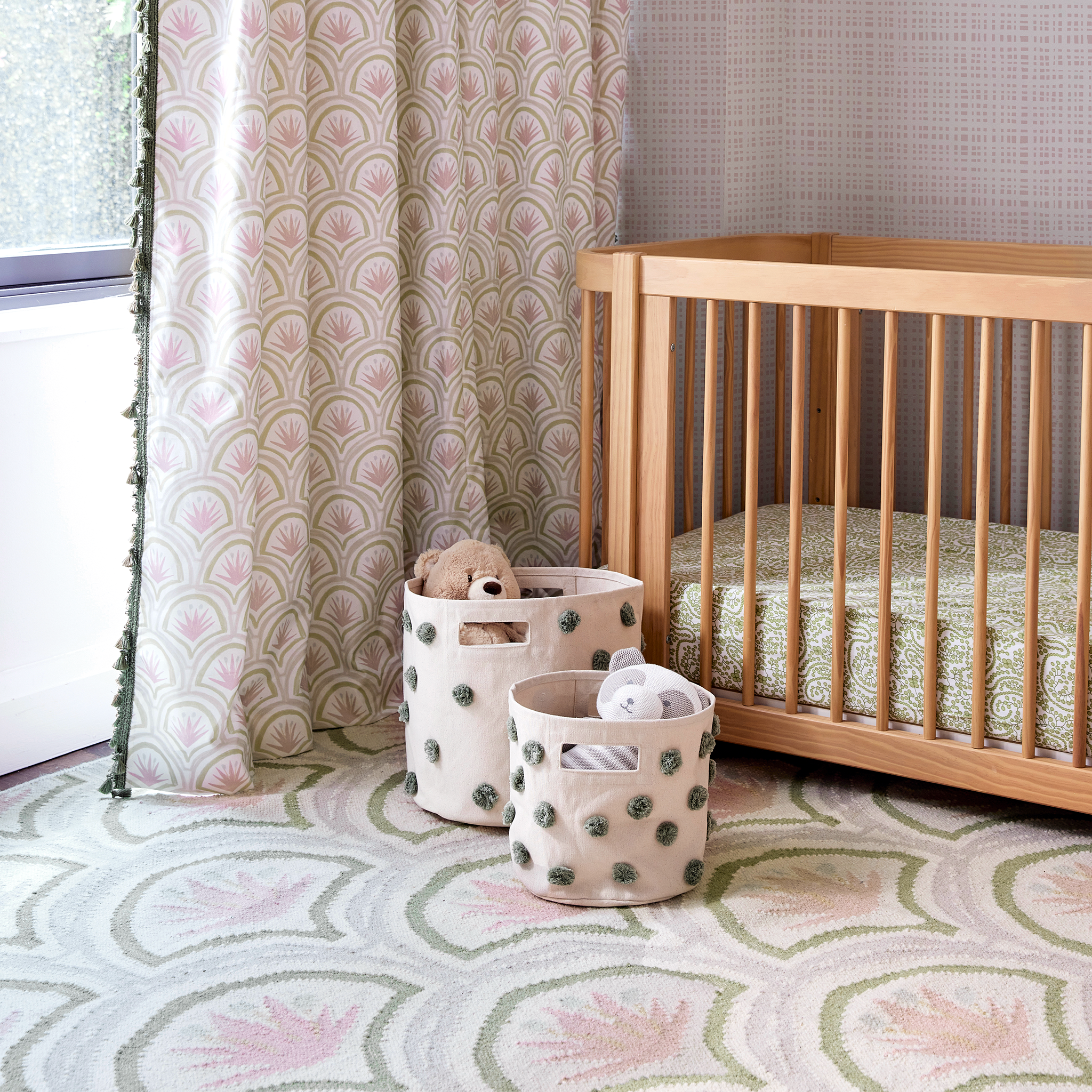 Pink and green art deco palm custom curtain hanging on a window next to a wooden crib and white toy baskets on a pink palm dhurrie rug