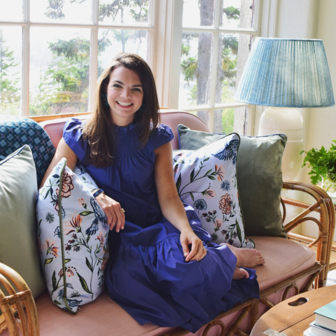 Brunette woman wearing a blue dress sitting on a pink couch in between two Daphne Powder custom pillows and two fern green velvet custom pillows