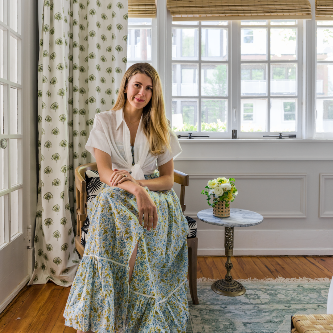 Blonde woman sitting on wooden armchair next to circular small table with white flowers in vase on top over illuminated window styled with Green Floral Custom Curtains