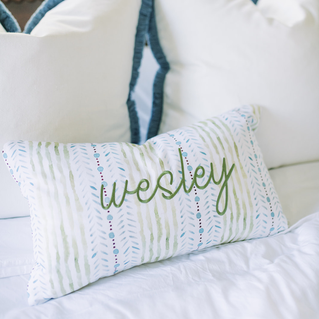blue and green striped pillow on a bed embroidered with "wesley" in front of white pillows with blue fringe