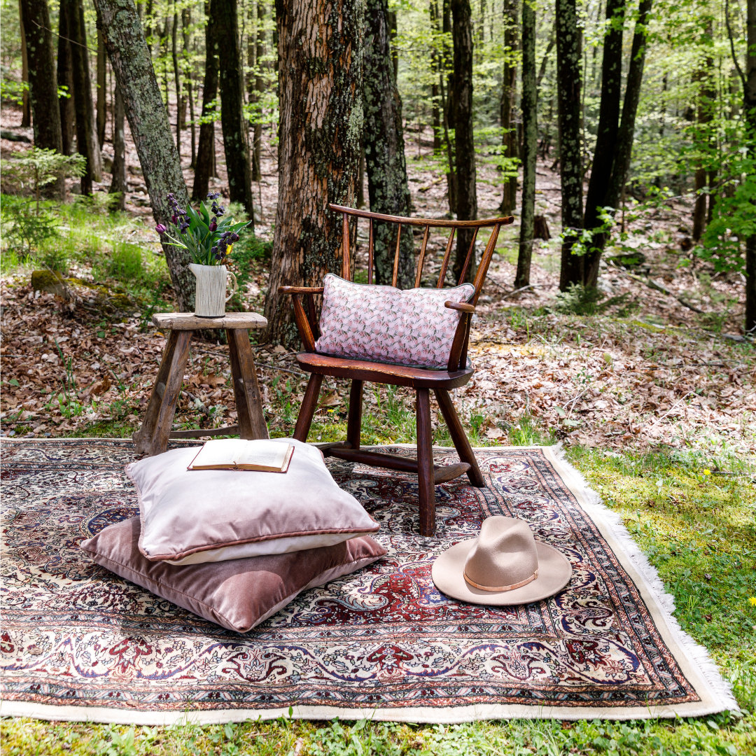 Printed rug laying in the forest with a stacked brown velvet custom pillow and a mauve velvet custom pillow and a book on top next to a wooden chair with a pink floral custom lumbar pillow